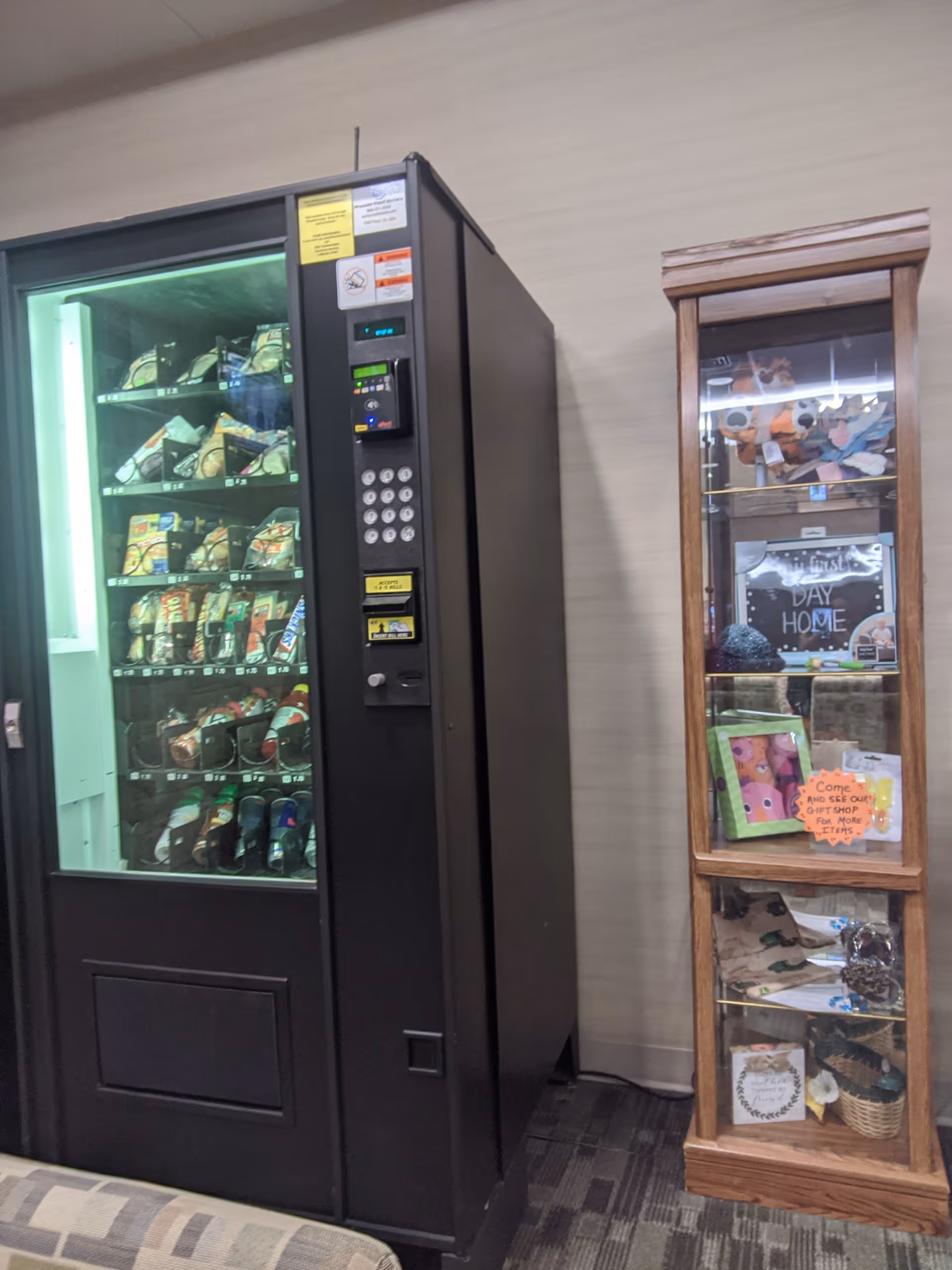 A tall black snack vending machine beside a wooden display cabinet in a hallway.