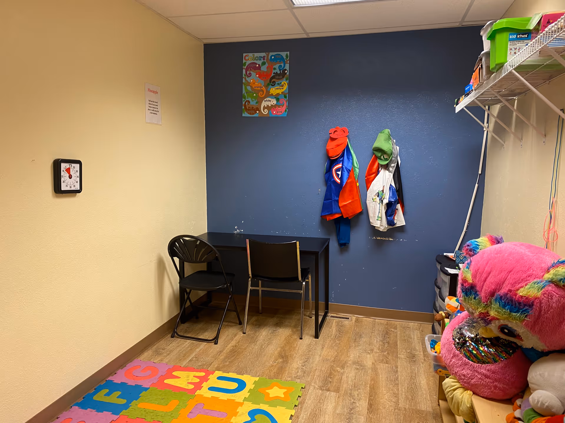 Small playroom with a blue accent wall, coats, desk, colorful foam mats, and stuffed toys.