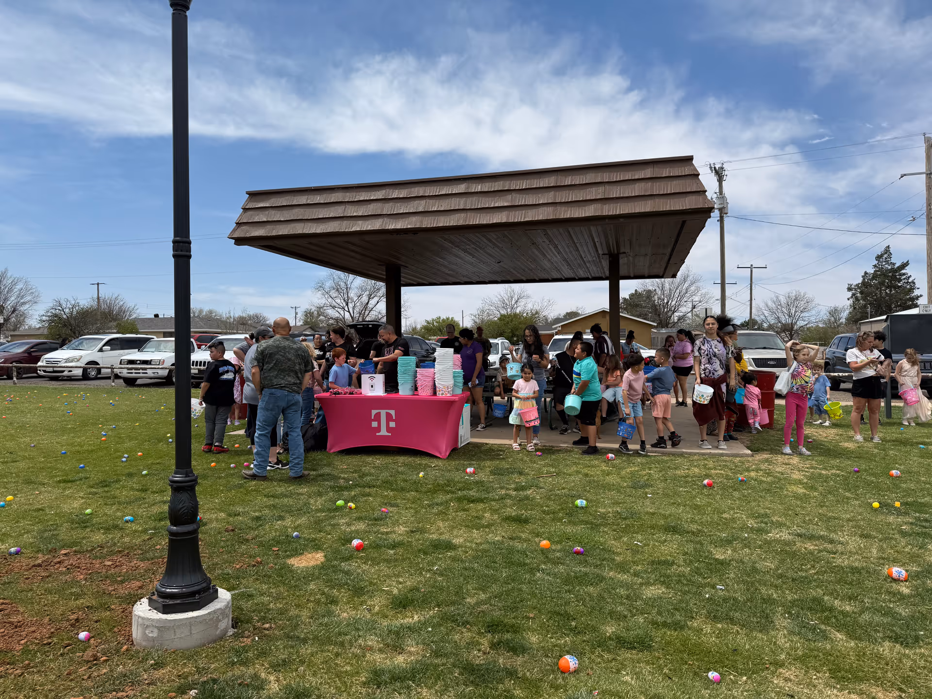 Outdoor gathering under a pavilion with many people and children around a pink table.