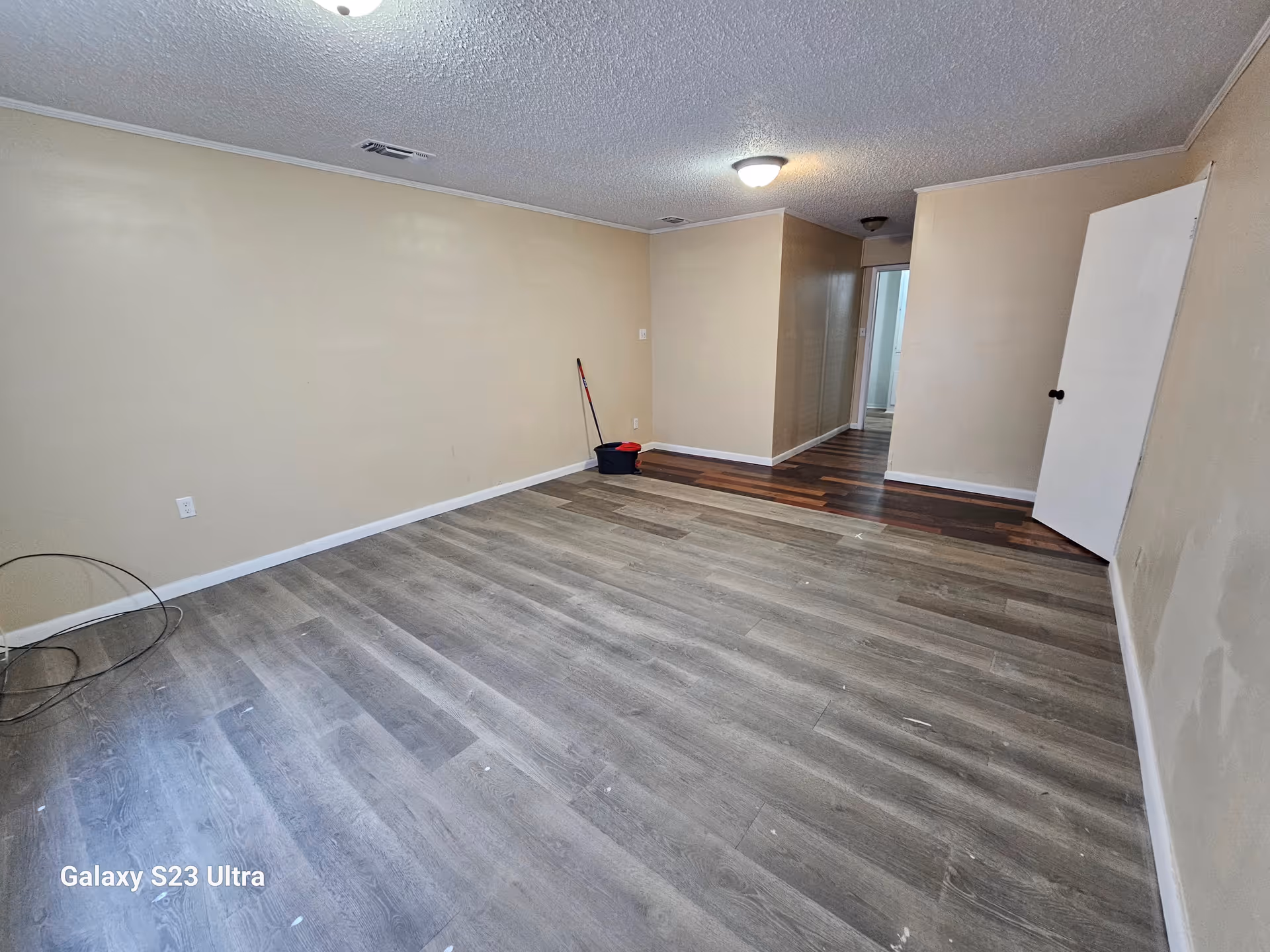 Empty living area with beige walls, mixed vinyl flooring, and a mop bucket in the hallway.