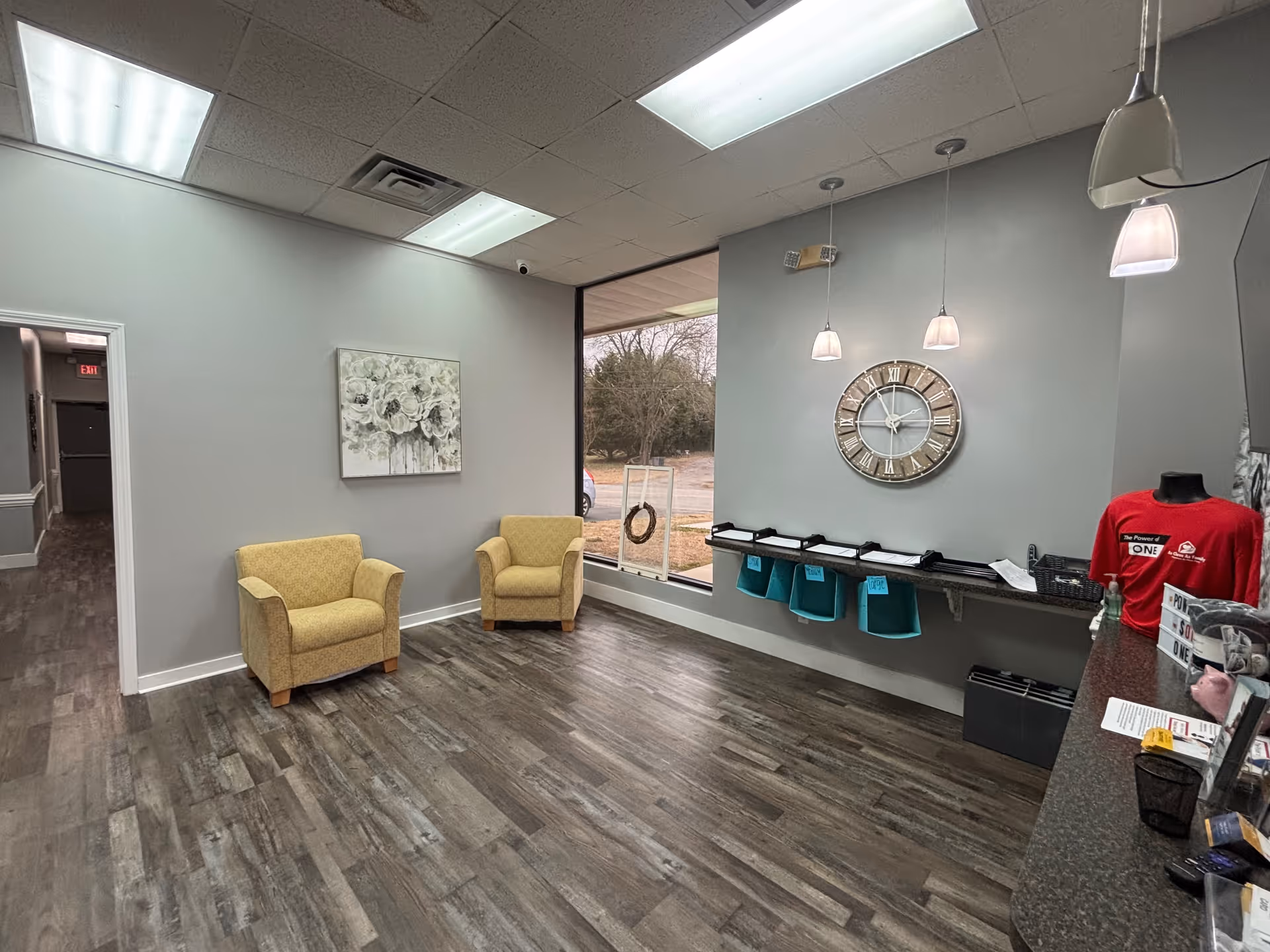 Interior reception area with two yellow chairs, wall clock, hanging lamps, and turquoise bins along counter.