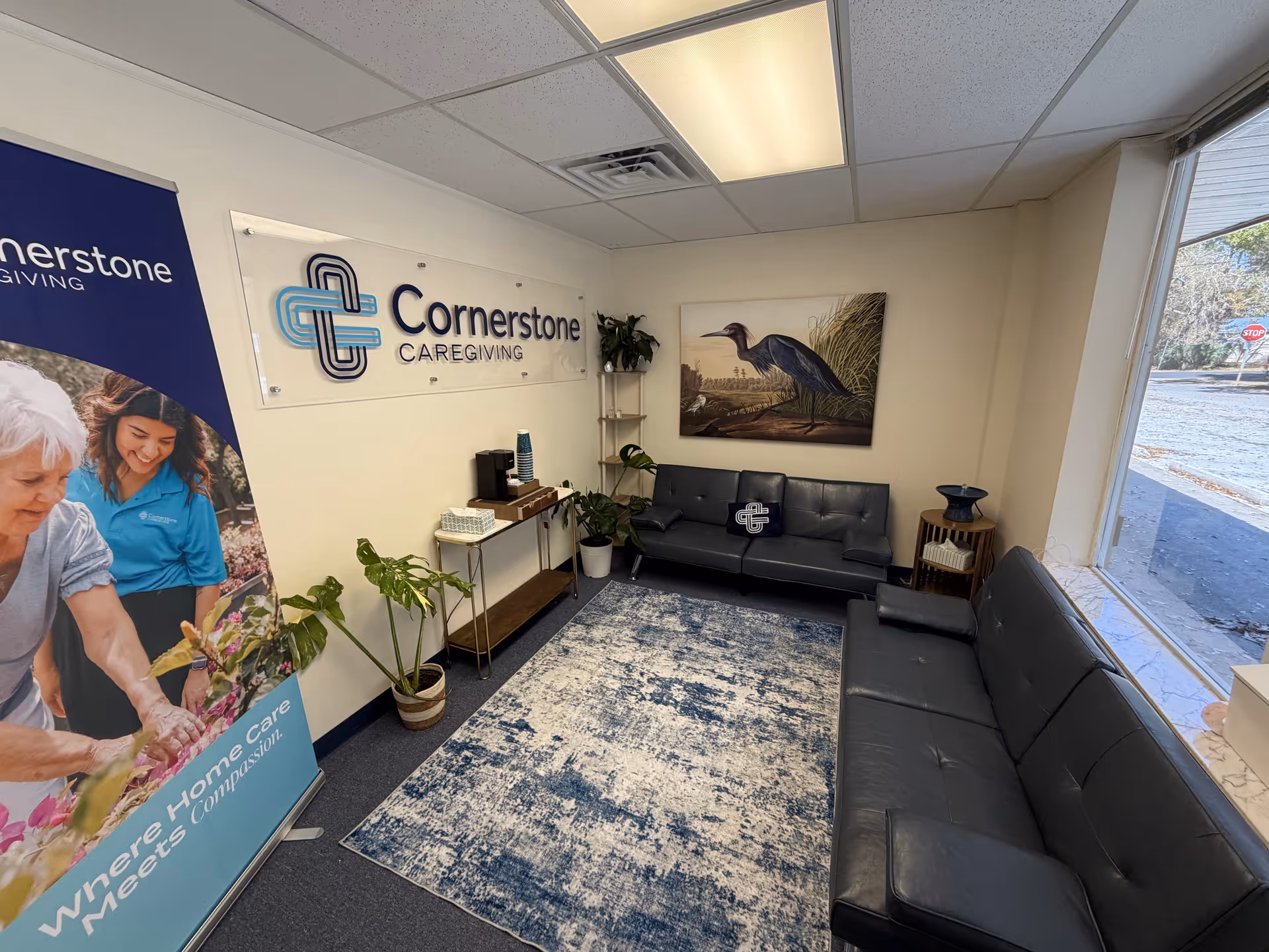 Interior reception area with sofa seating, a Cornerstone Caregiving sign, plants, and artwork.