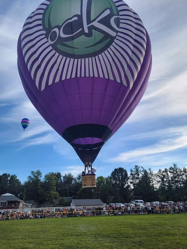 Large purple hot air balloon ascends over a crowd at a grassy field.