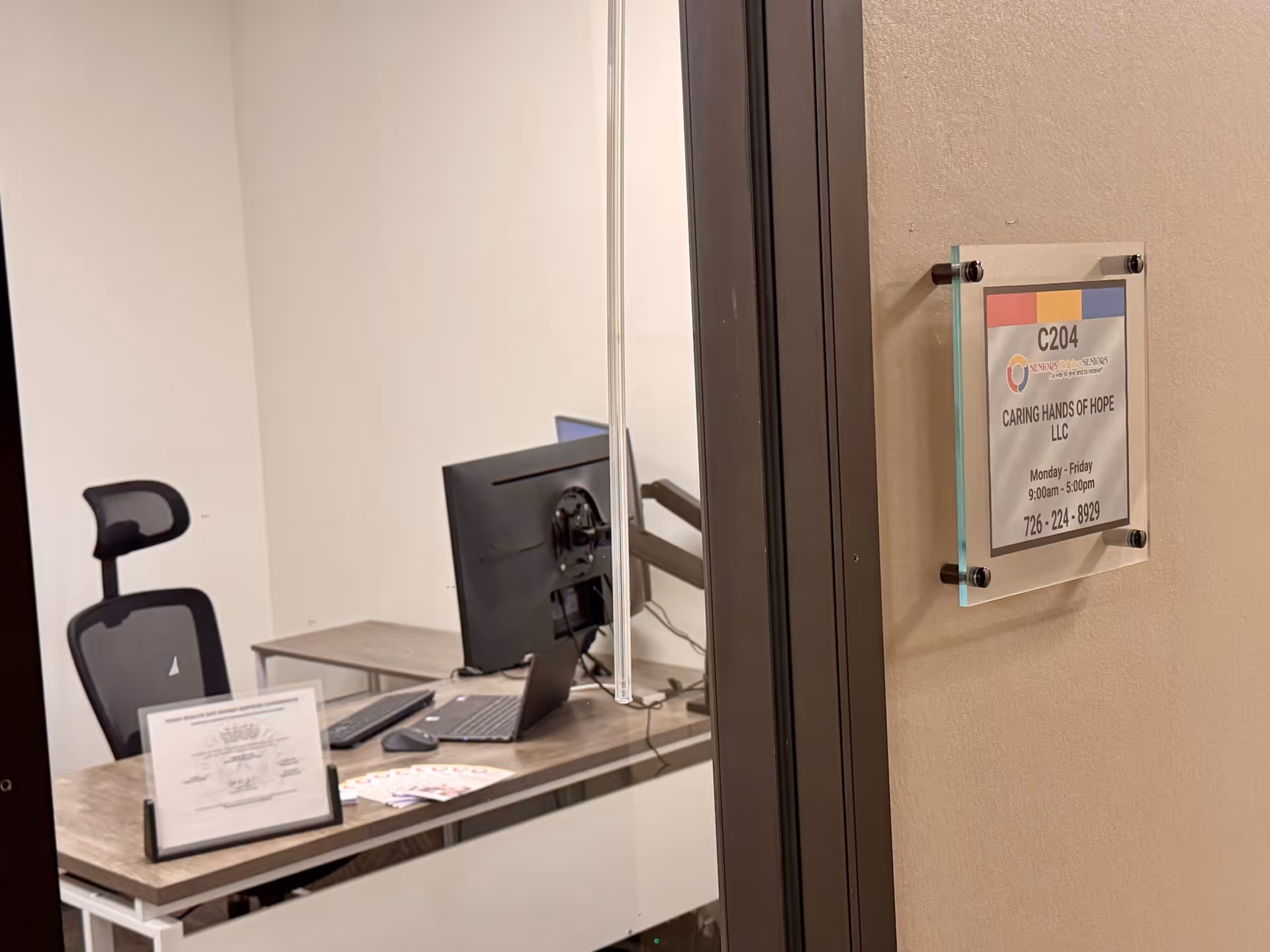 Office desk with computer and chair inside a glass-walled room, sign on adjacent wall.