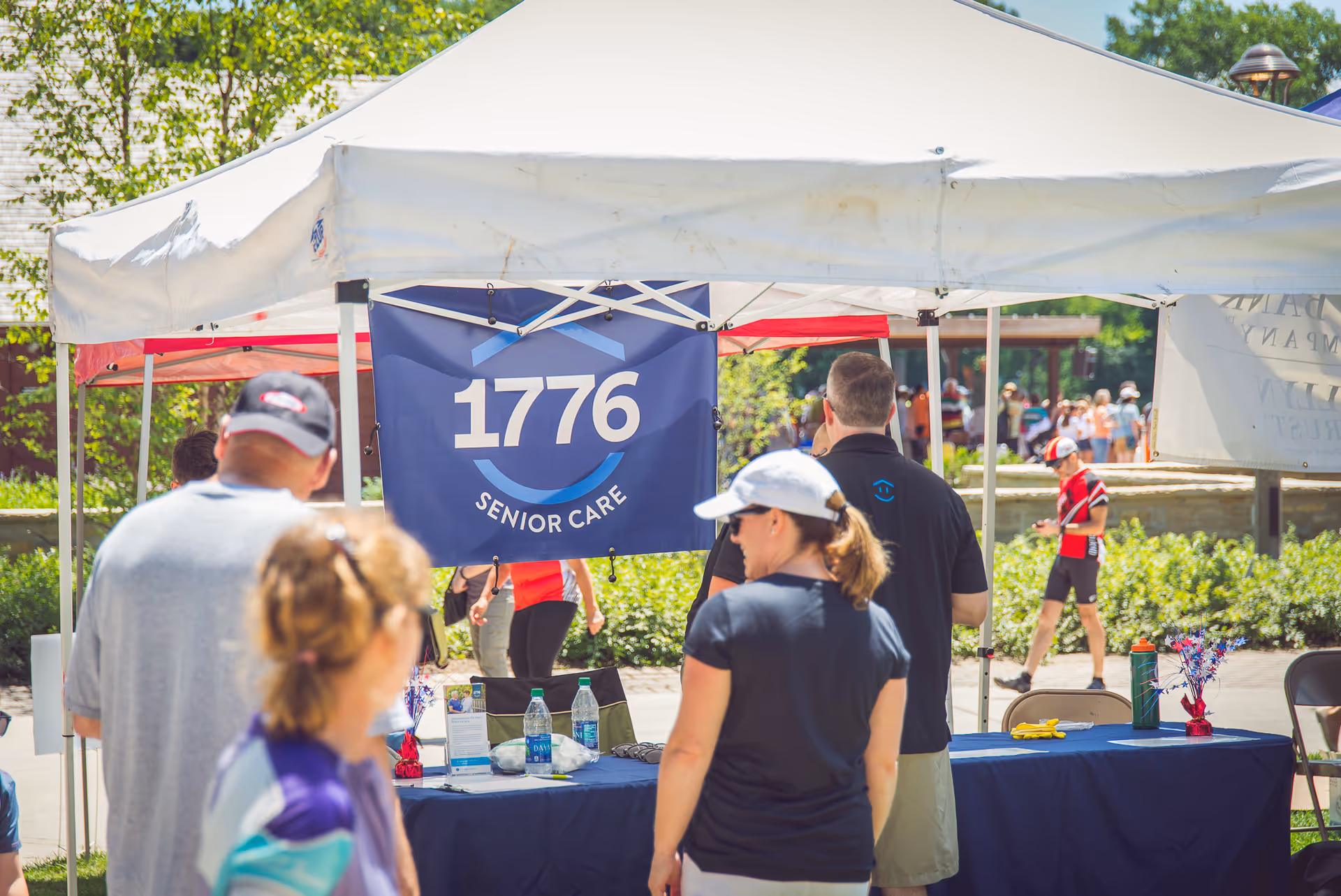 Outdoor event at 1776 Senior Care with a banner tent and attendees around a table.
