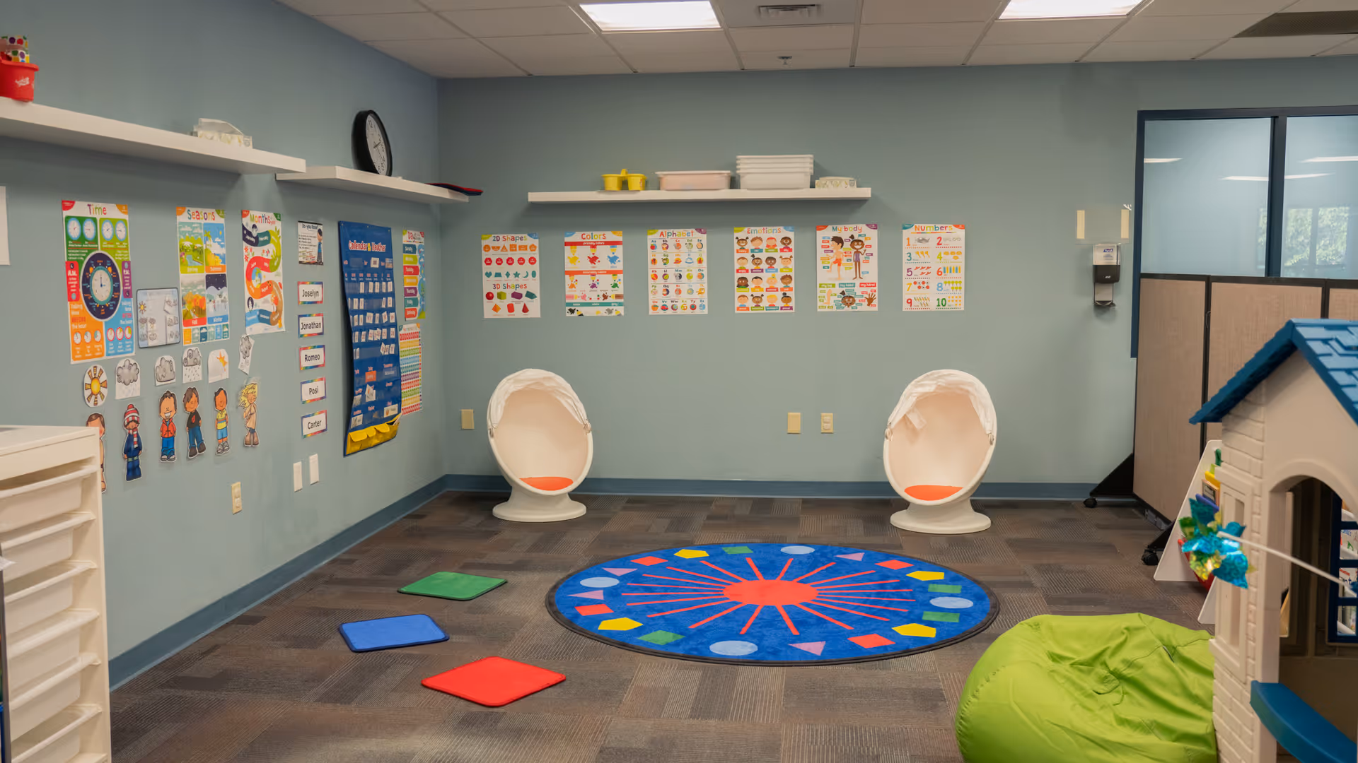 Interior playroom for residents with colorful rug, posters, two white circular chairs, and a toy house.