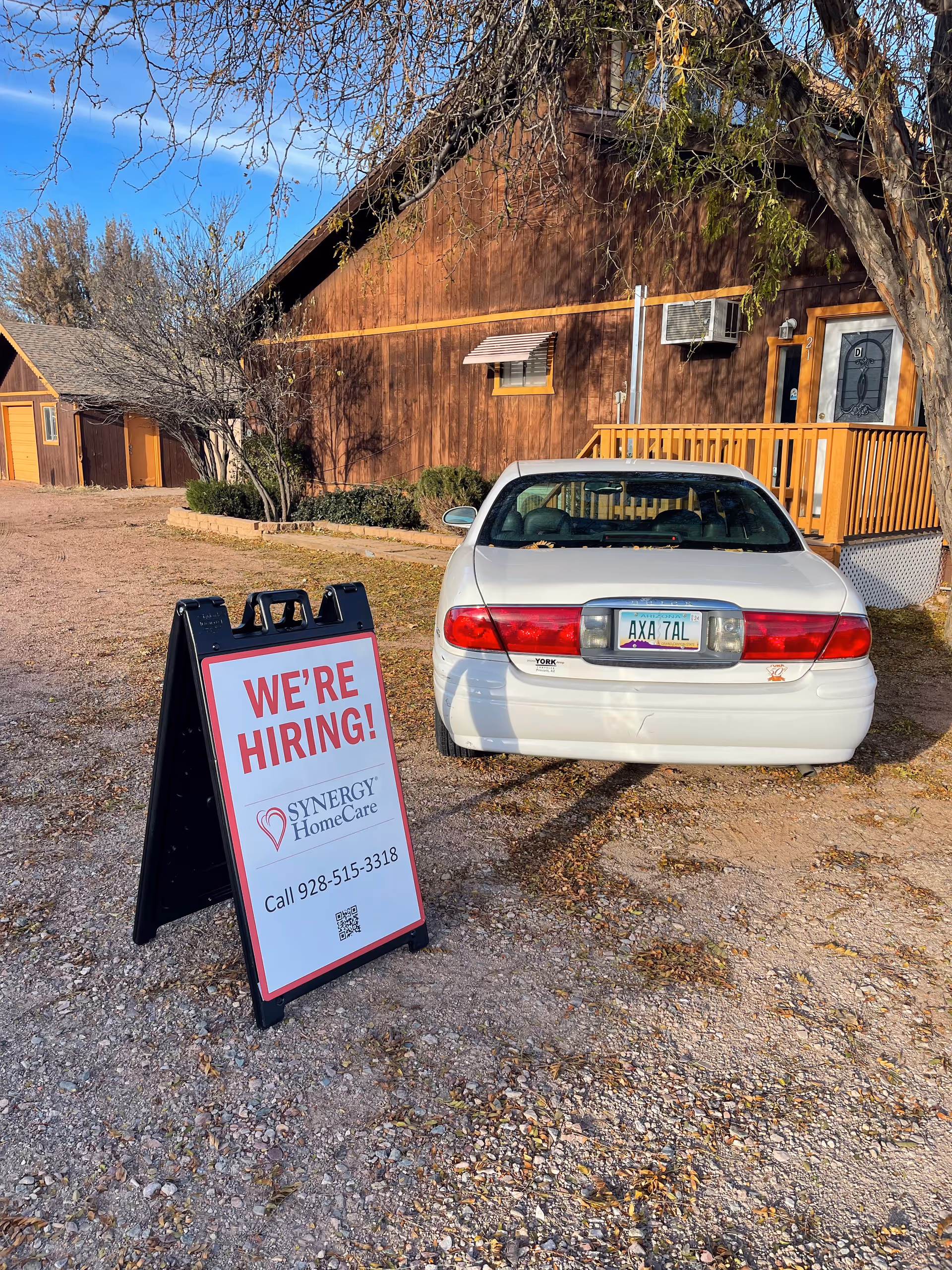 Exterior view of a brown Synergy HomeCare building with a 'We're Hiring' sign and a car.
