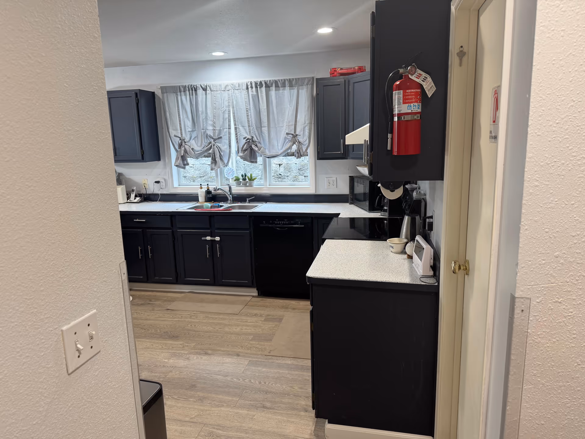 Narrow kitchen with dark cabinets, light countertops, window with curtains, and a wall-mounted fire extinguisher.