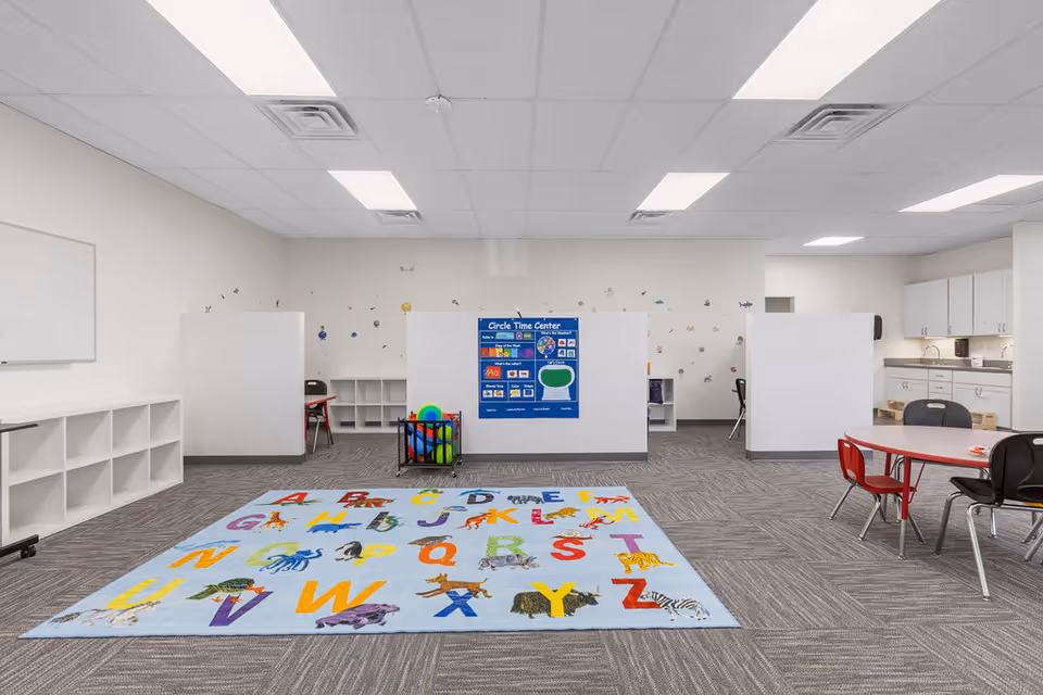 Bright classroom-style activity room with alphabet rug, cubbies, partitions, and a kitchenette in back.