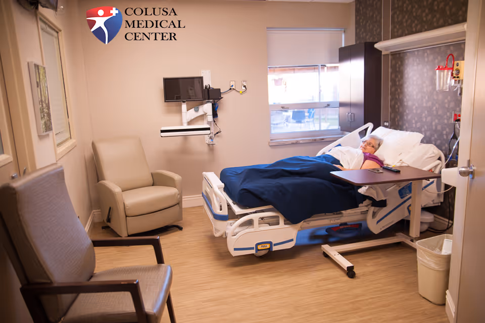 Hospital room with patient in bed, chairs, medical equipment, and Colusa Medical Center sign.