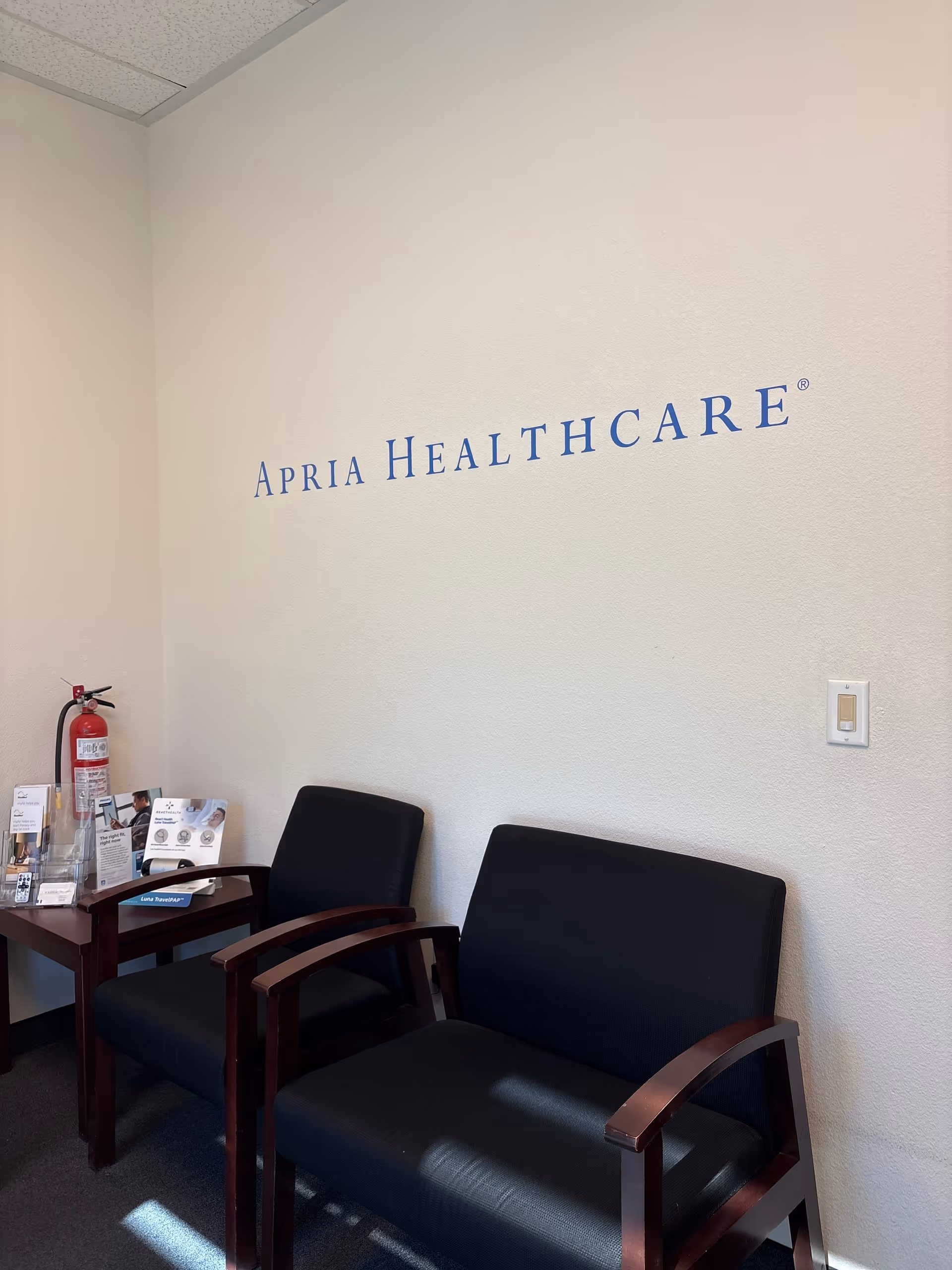 Interior waiting area with two black chairs, brochures on the table, and Apria Healthcare logo.