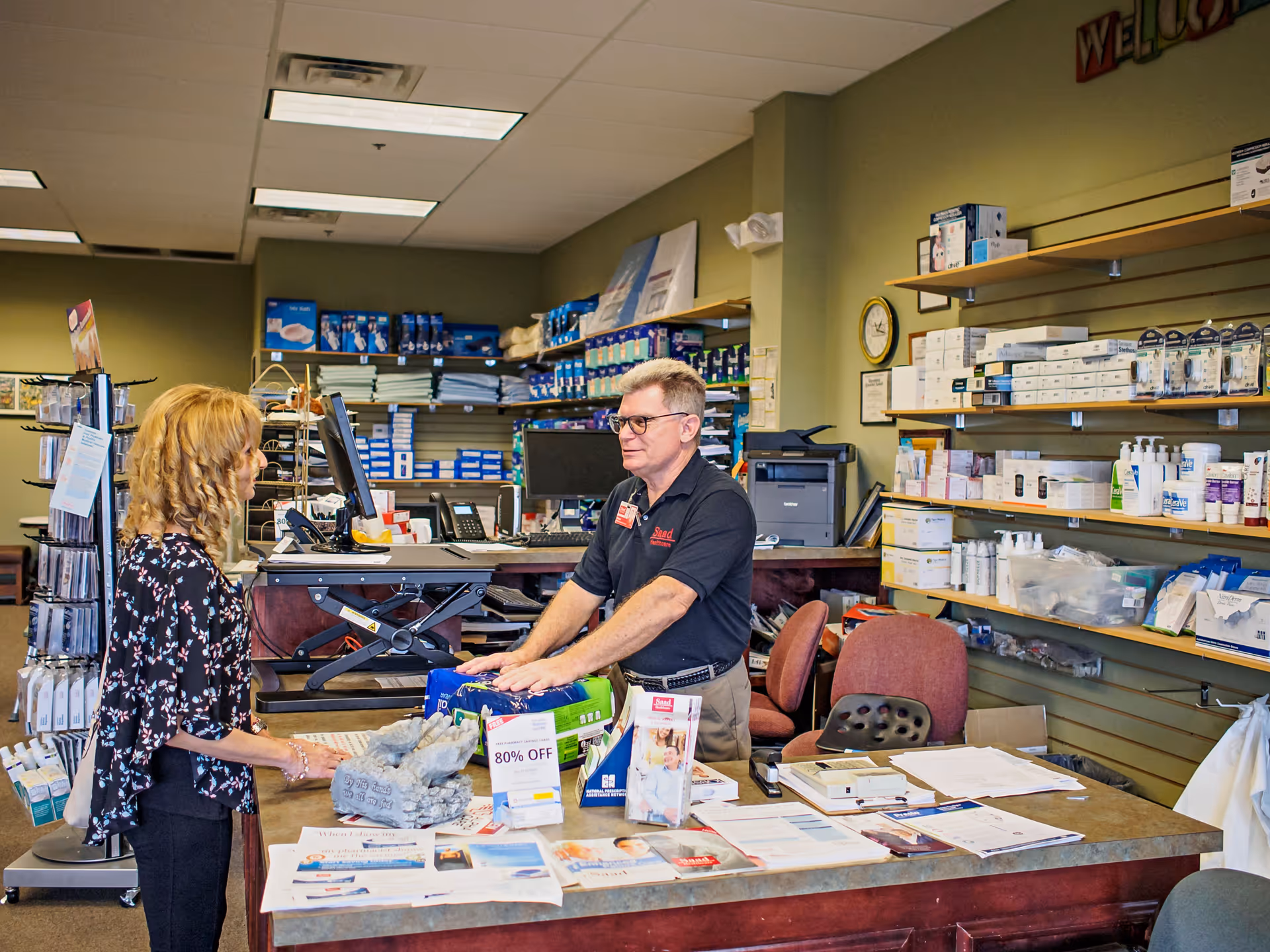 A man behind a checkout counter assists a woman in a well-stocked interior pharmacy.
