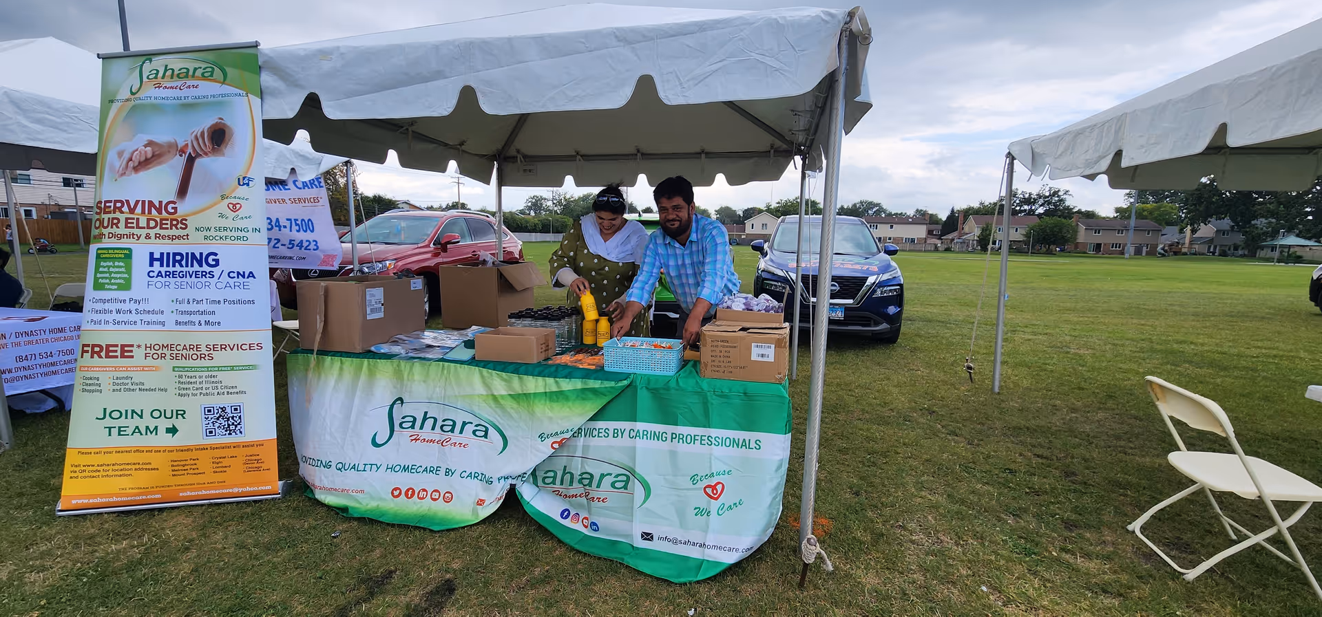 Outdoor Sahara Home Care booth under tents with two staff arranging boxes on a table.