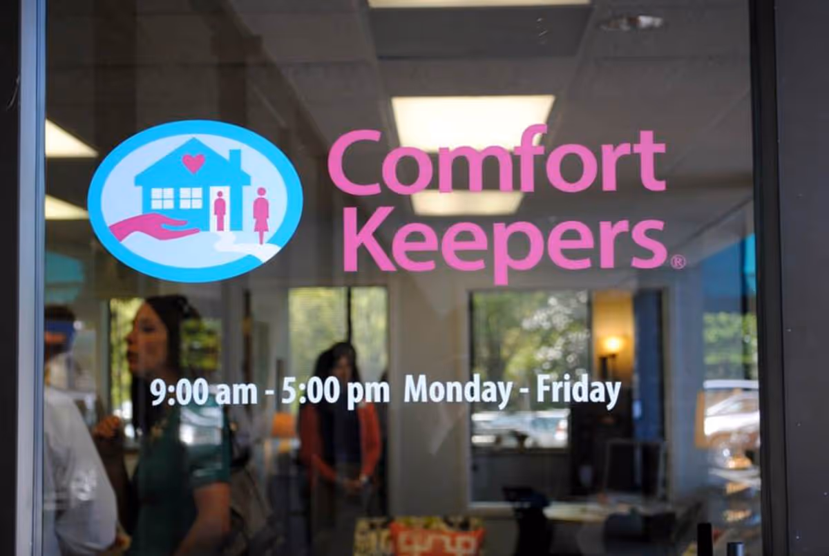 Glass storefront displays Comfort Keepers logo and hours with people visible inside.