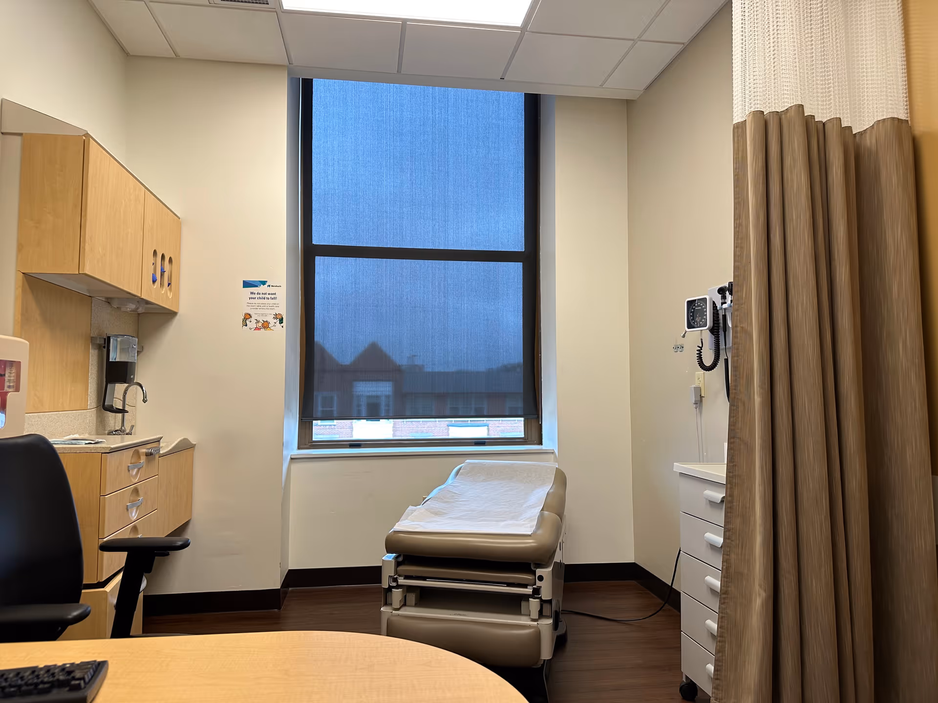 Medical examination room with a chair, cabinets, sink, and a window with blue blinds.