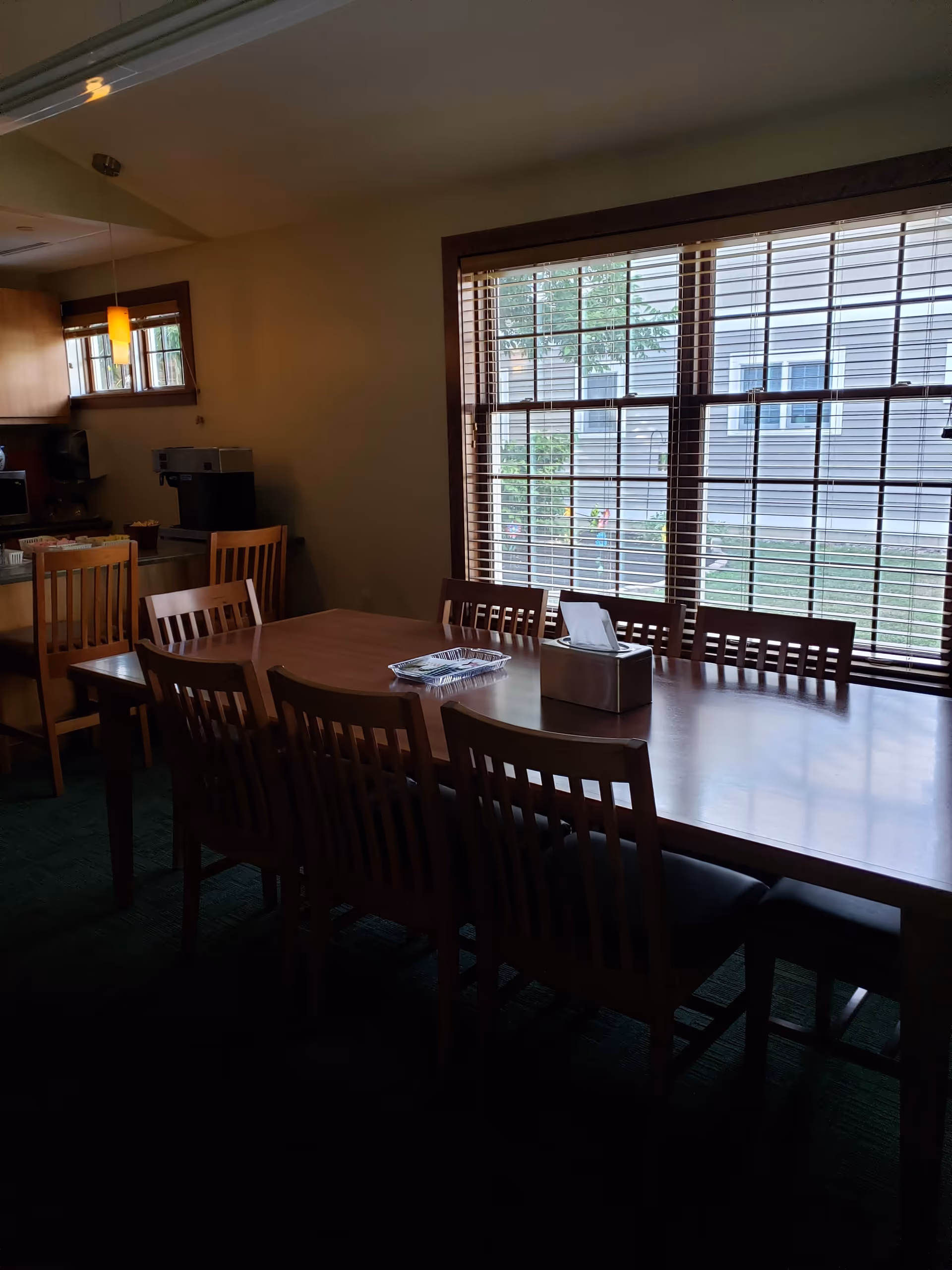 Long wooden dining table with chairs in a bright dining area by large windows.