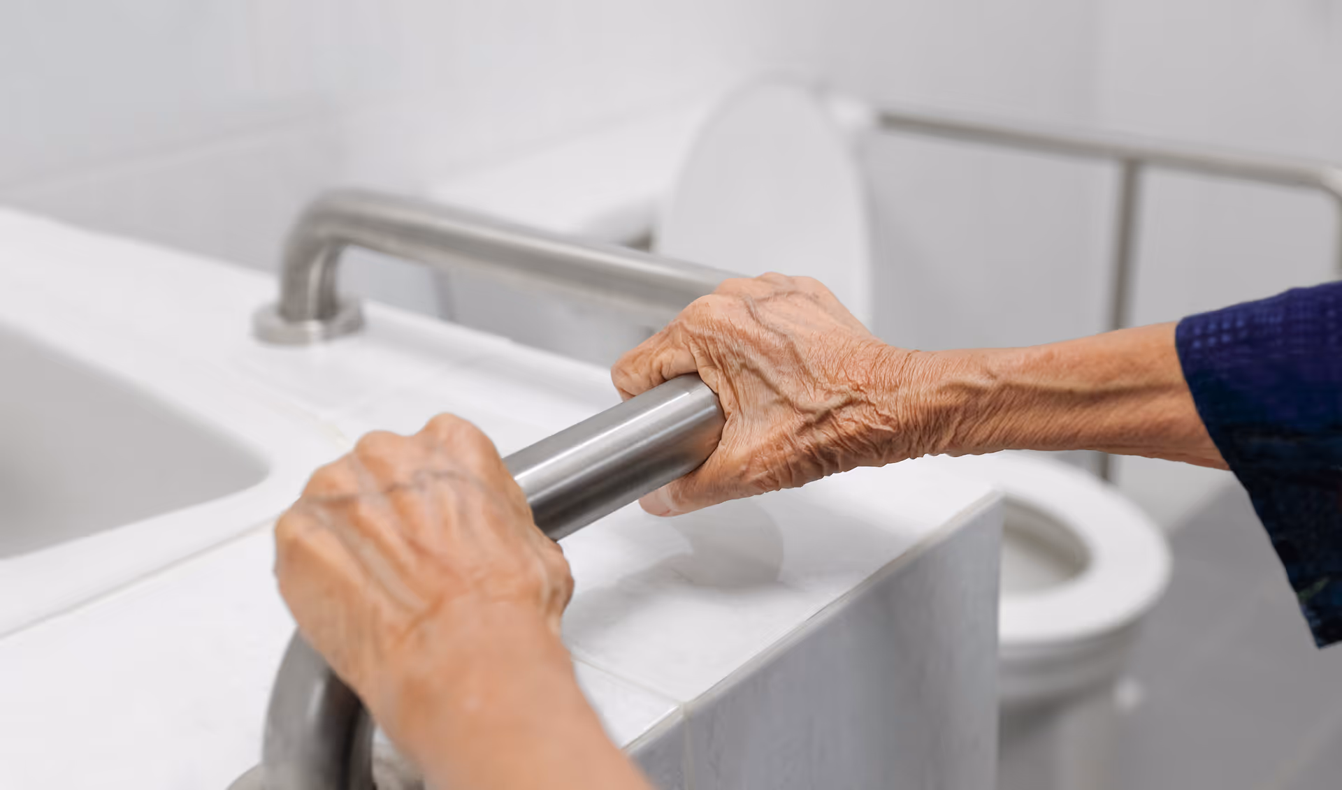 Close-up of elderly hands gripping a metal grab bar beside a bathroom sink.