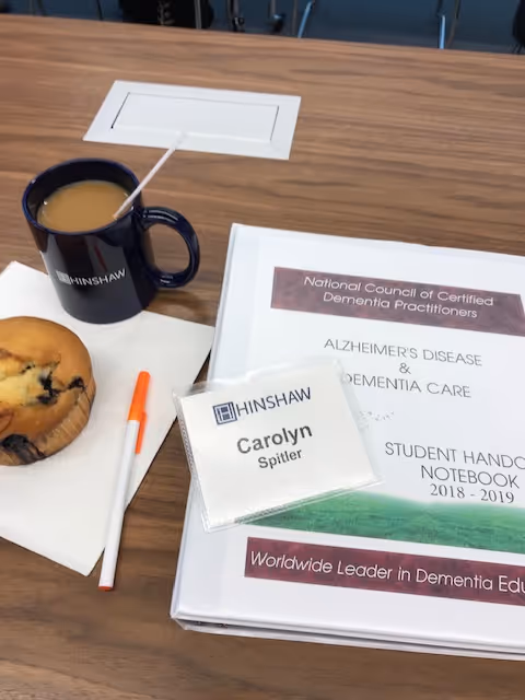 A table with a muffin, coffee mug, name badge, and dementia care binder.