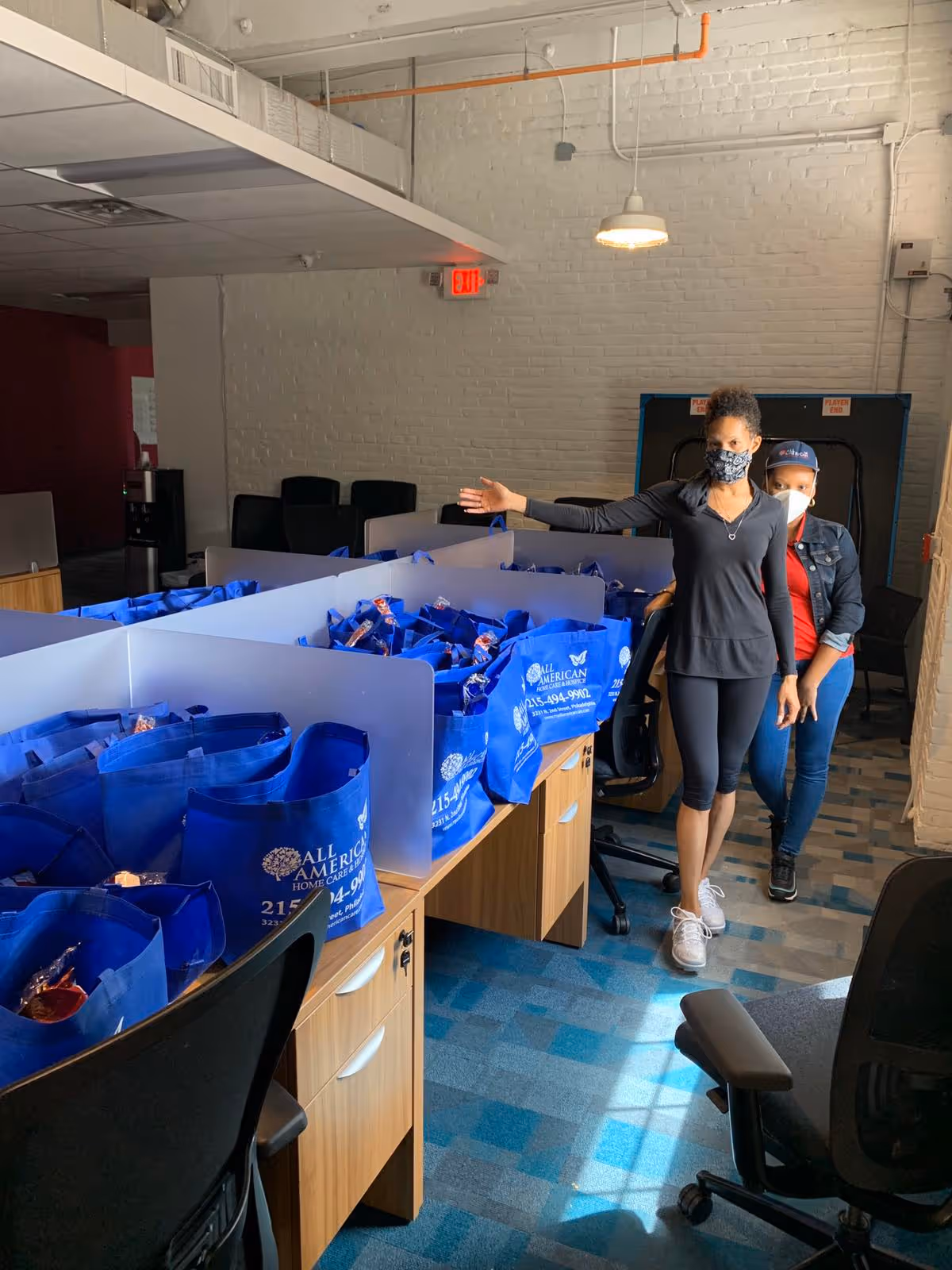 Two masked women stand beside blue All American Home Care tote bags in an office workspace.