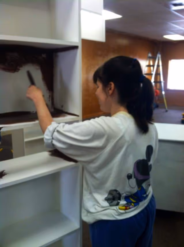 Person painting a cabinet inside a facility interior with ladders visible in background