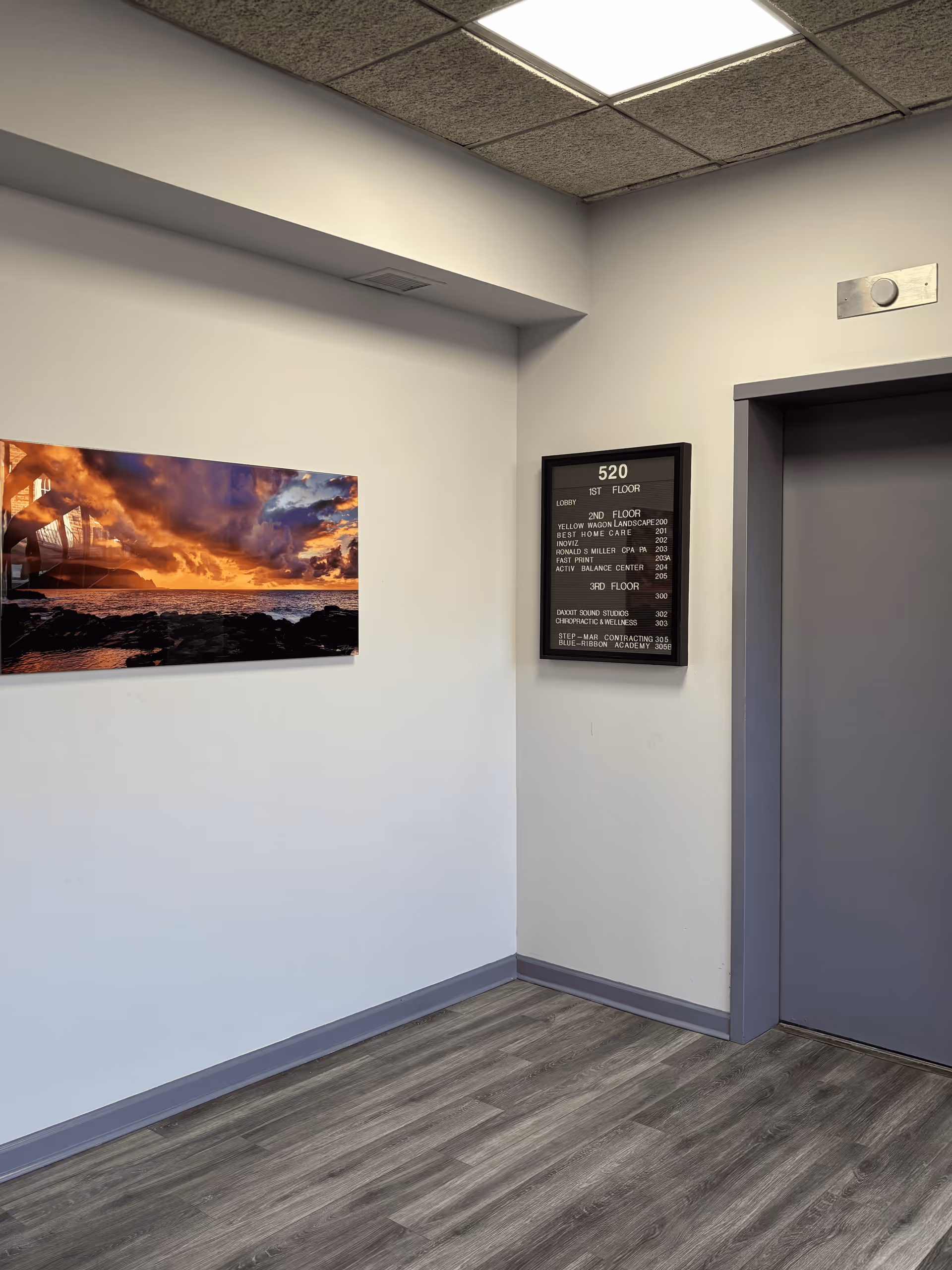 An empty hallway interior with a framed sunset photograph and a directory sign near a door.