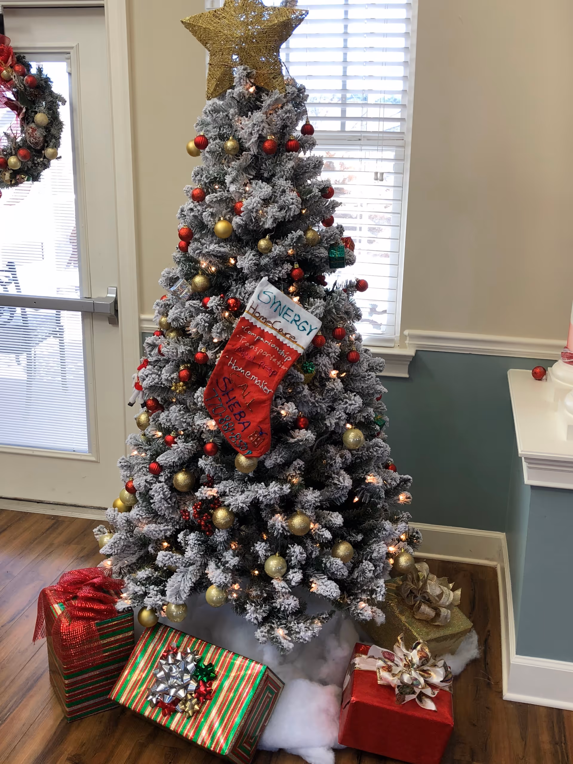 Snow-dusted Christmas tree with presents in a decorated indoor facility lobby.