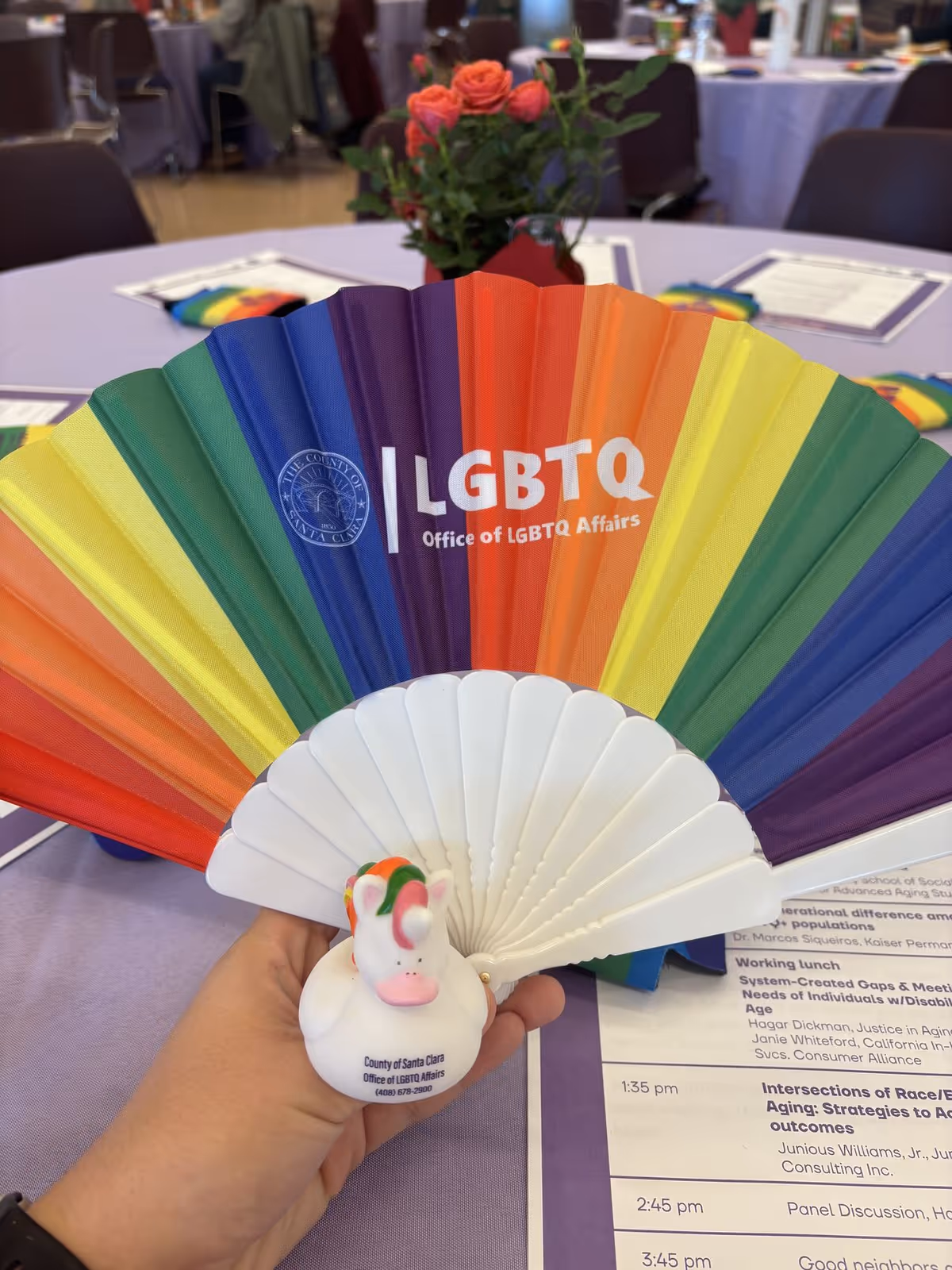 Hand holds rainbow LGBTQ fan in foreground; banquet tables and flowers blurred in background.