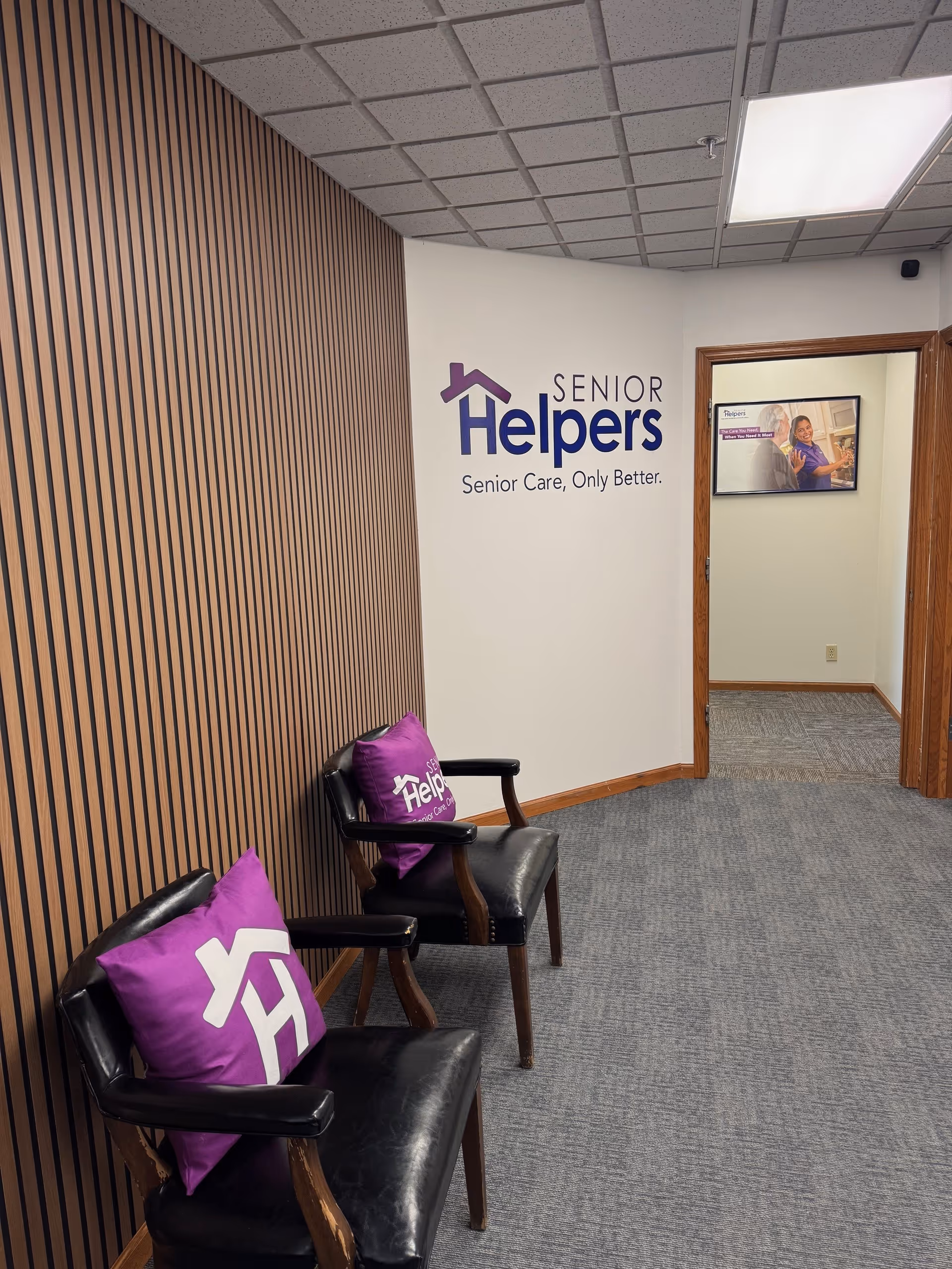 Hallway reception area with striped wood wall, black chairs, and Senior Helpers logo.