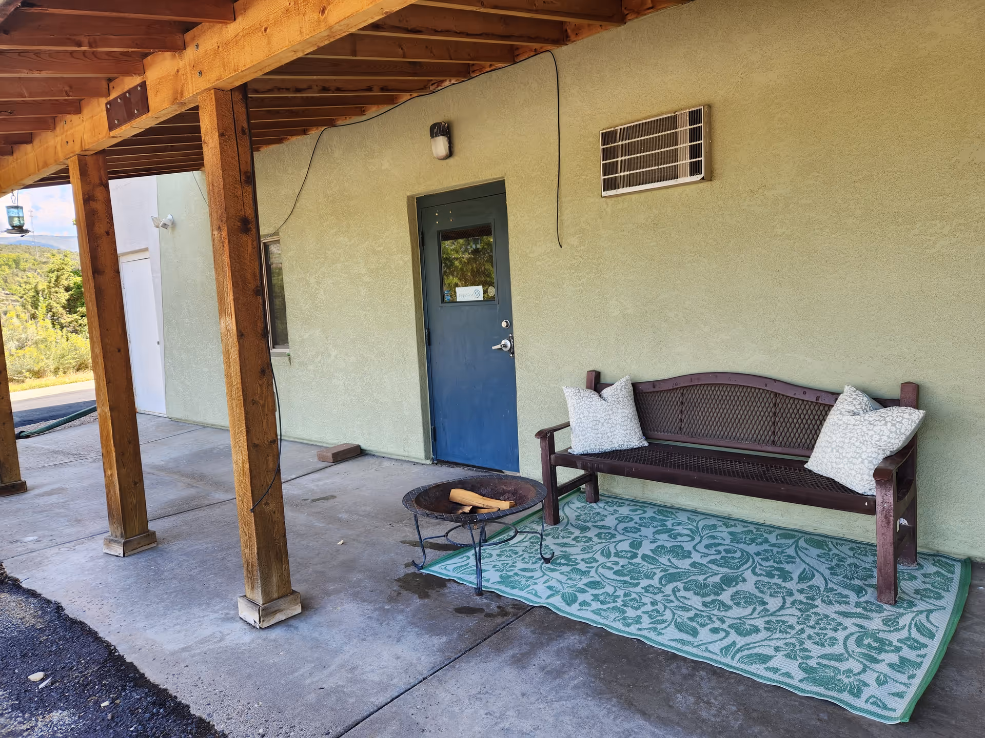 Exterior covered patio with wooden posts, blue door, bench, cushions, and patterned rug.