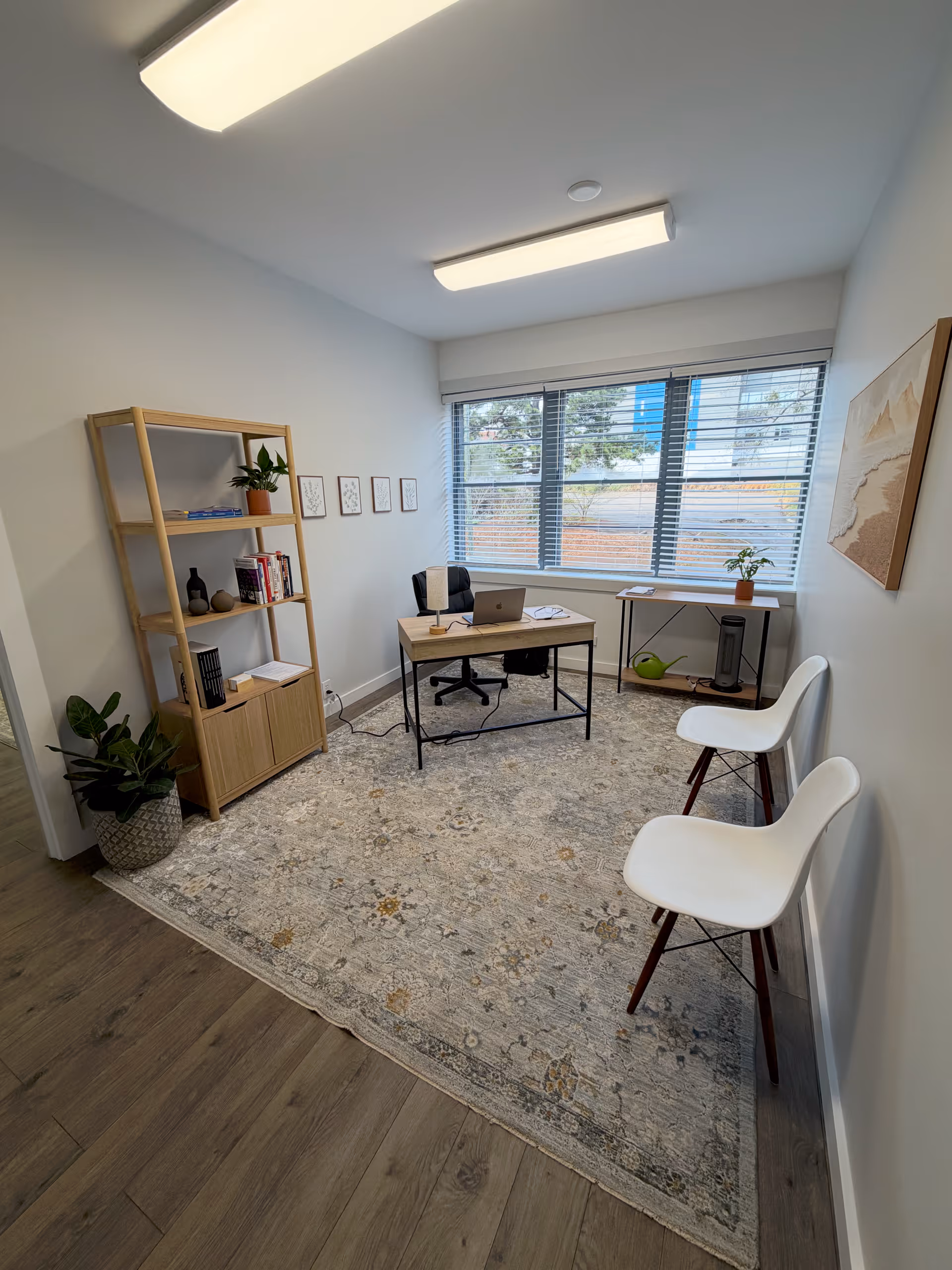 Small modern office area with desk, laptop, shelves, two white chairs, and a large window.