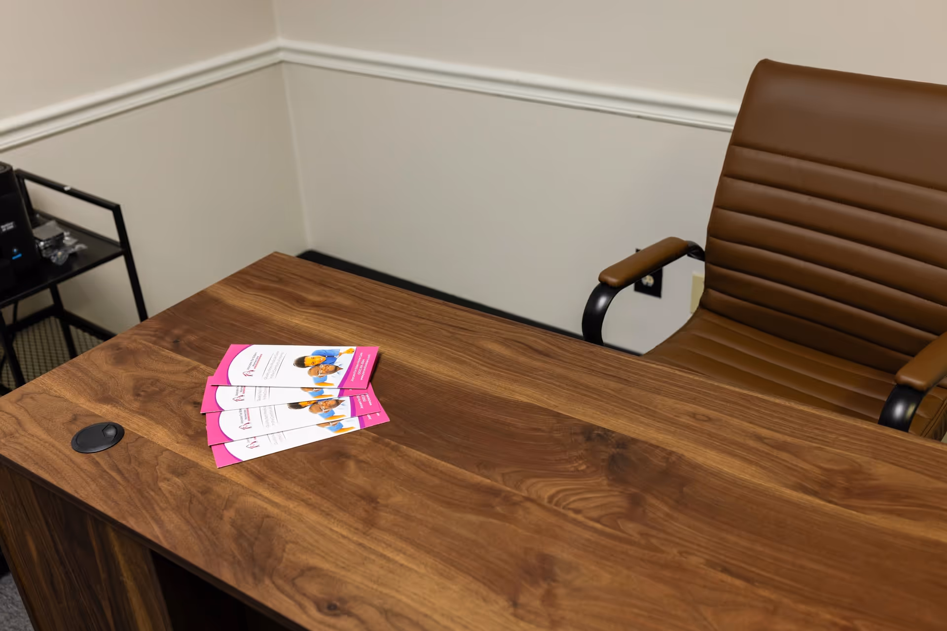 In a small office, a wooden desk with brochures and a brown leather chair.