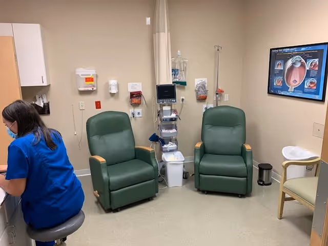Medical treatment room with two green recliner chairs, medical equipment, and a nurse at a desk.