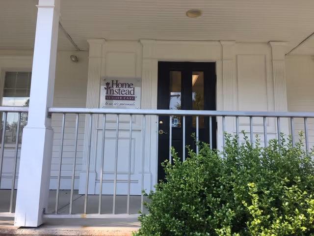 White front porch of Home Instead Senior Care with a sign beside the door and shrubs.