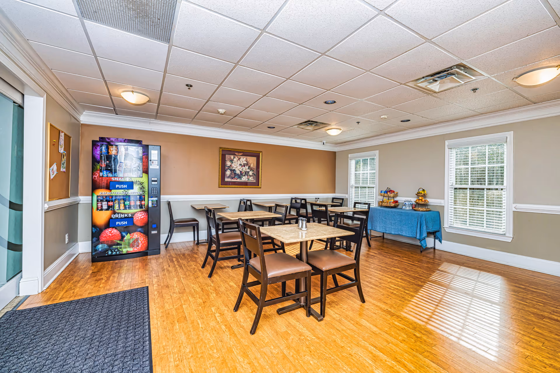 Bright dining room with wooden tables, chairs, a vending machine, and a snack table.
