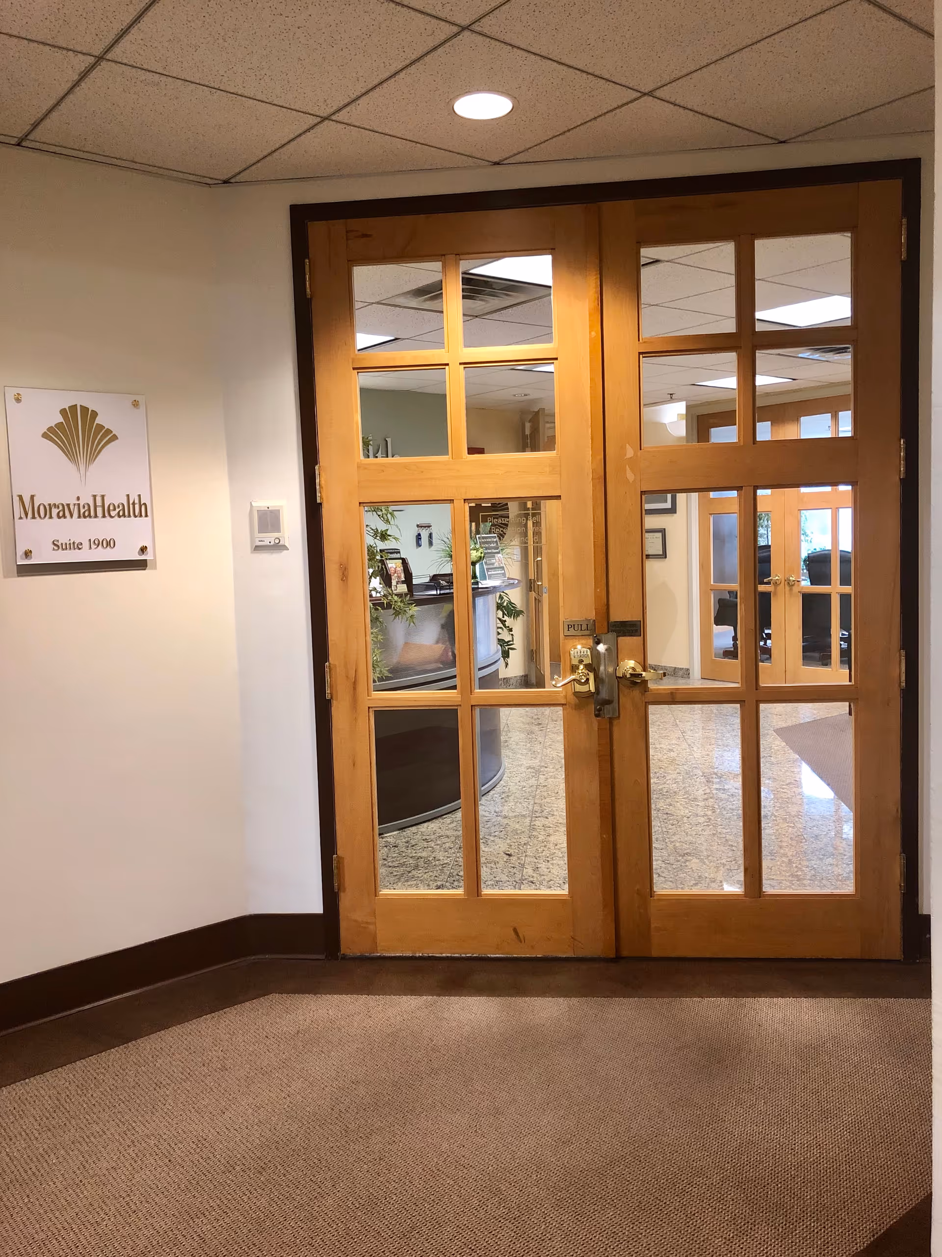 Wooden glass double doors open into a MoraviaHealth interior lobby with a carpeted floor.