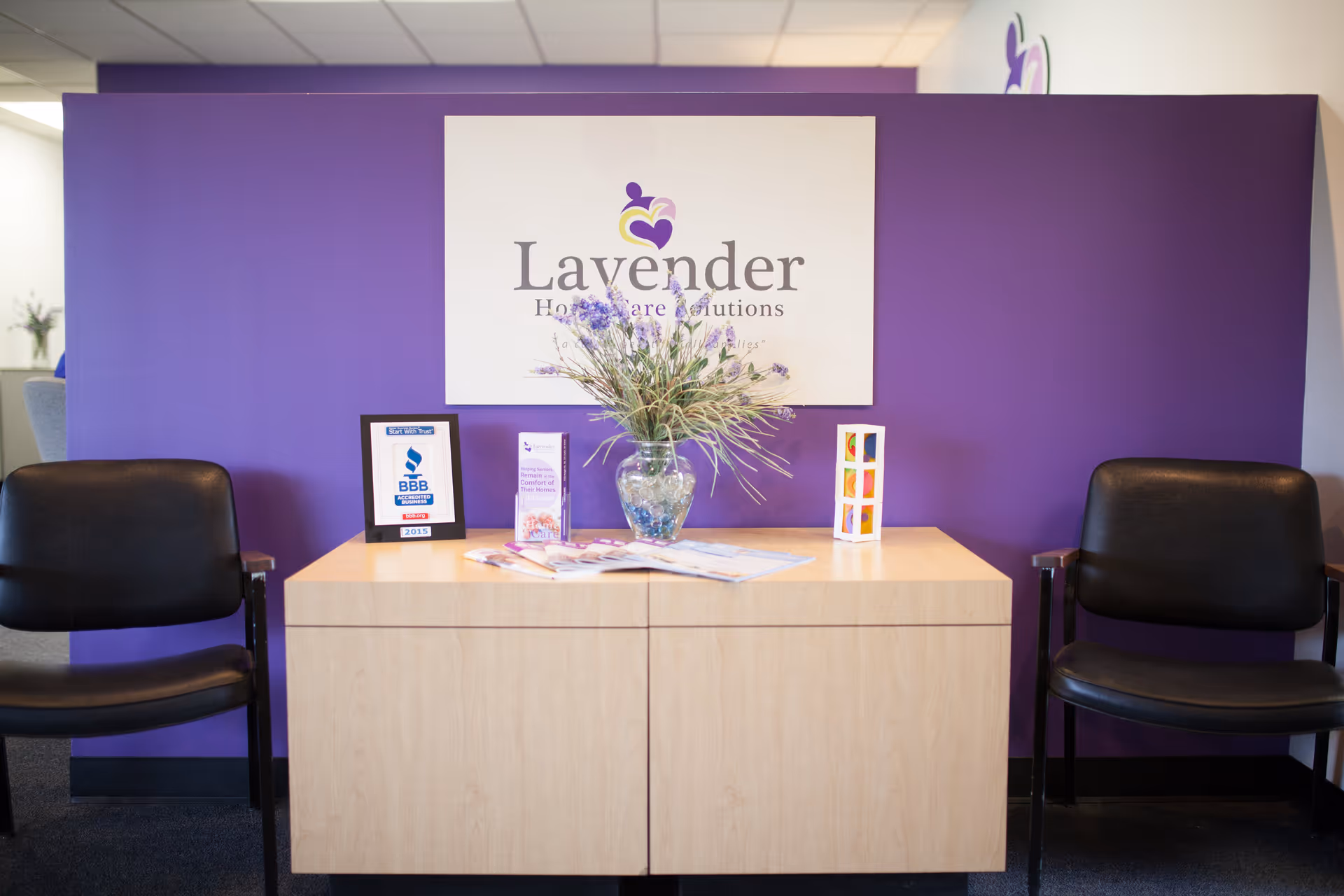Reception area with lavender wall, sign, vase, and two black chairs.