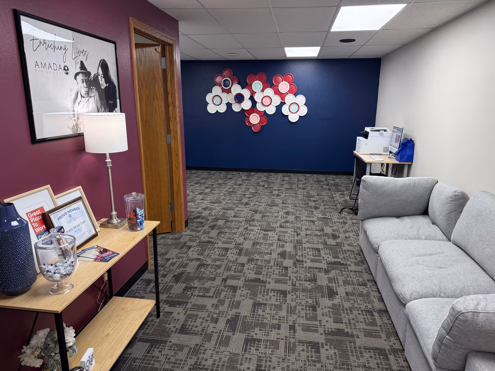 Interior waiting area with purple accent wall, gray sofa, and pink-white floral wall art.