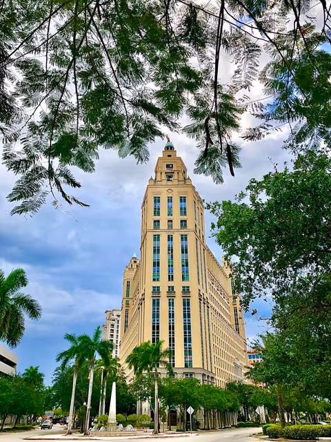 Tall beige skyscraper-style building framed by green trees and blue sky.