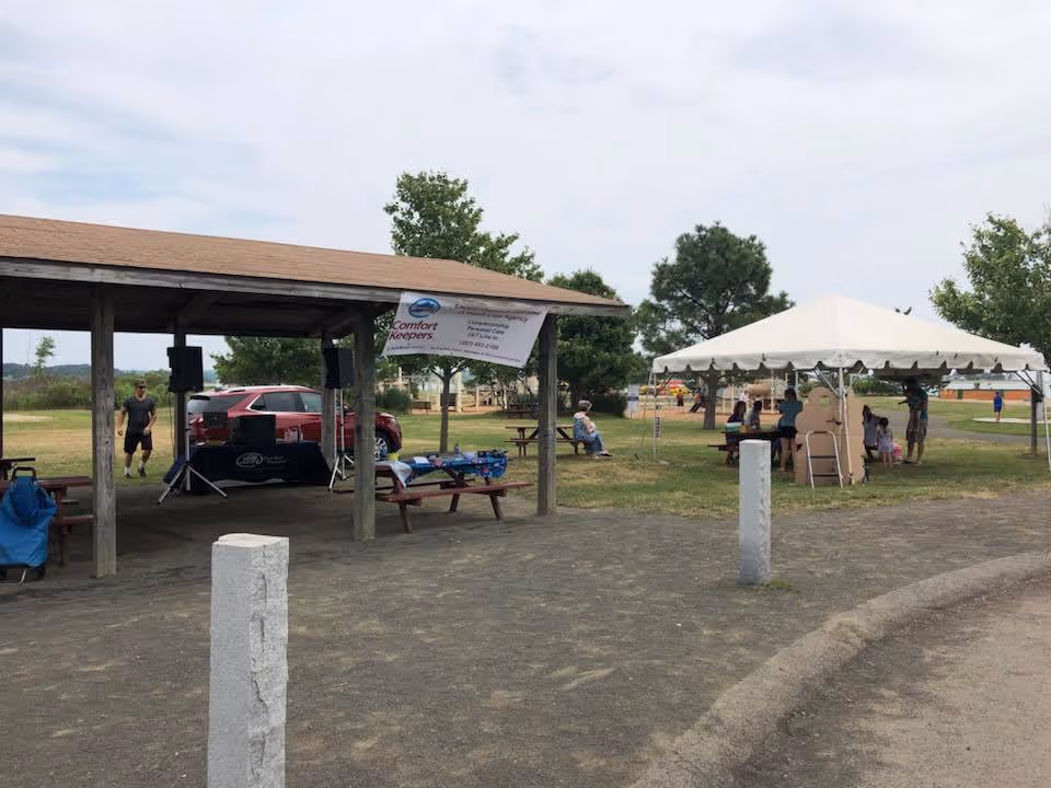 Outdoor park area with pavilion, banner reading Comfort Keepers, tent, and people at an event.
