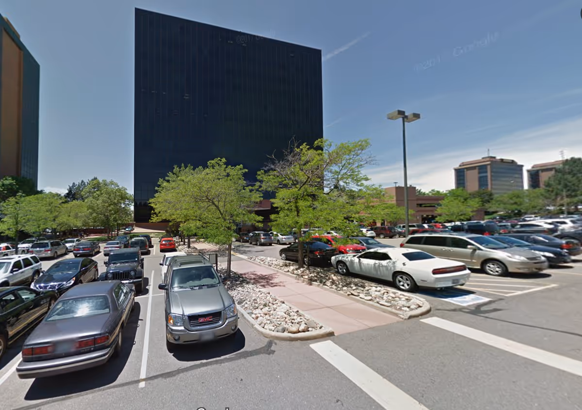 Parking lot in front of a tall dark glass building with trees and cars.