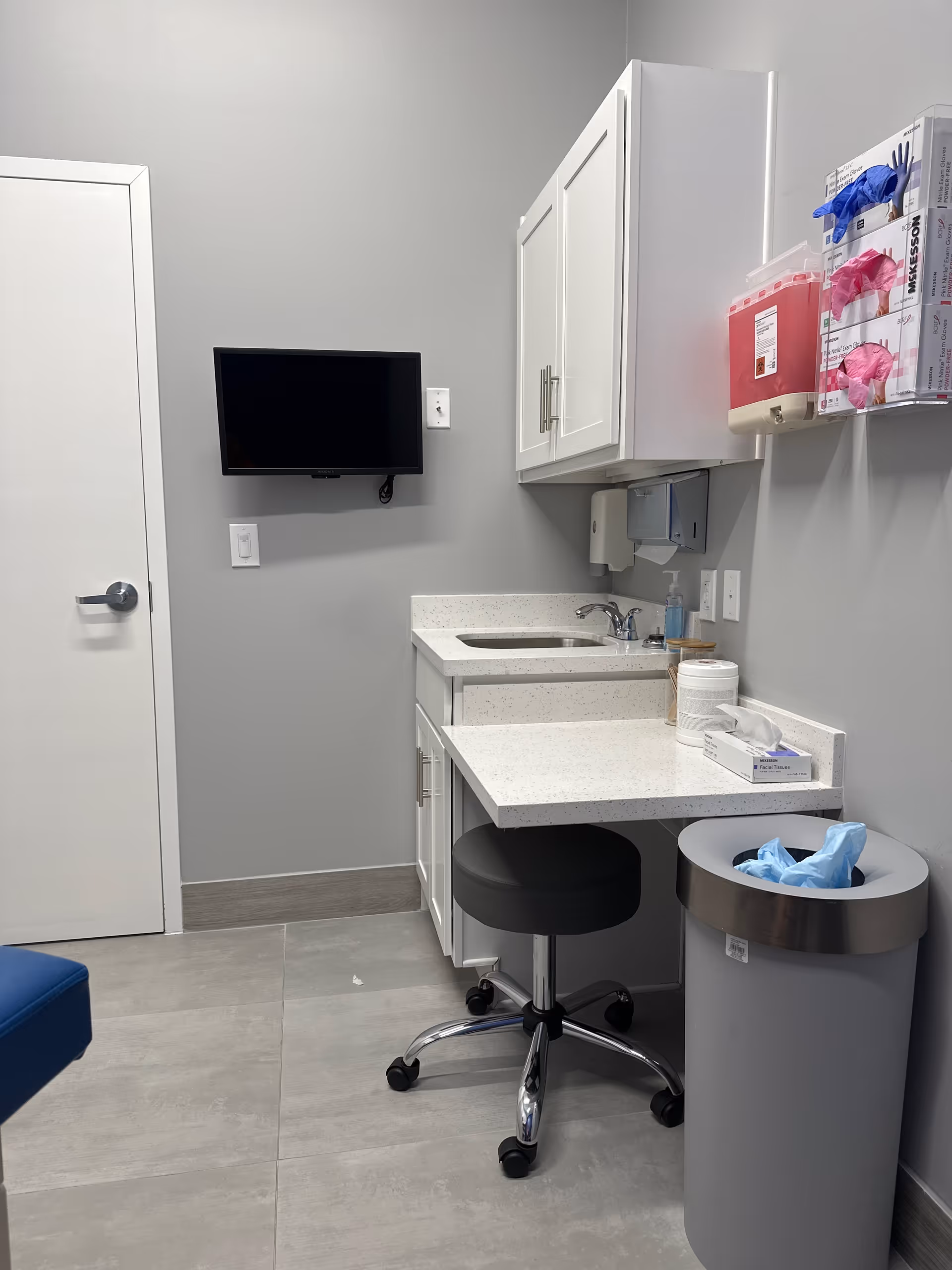 Small clinical kitchenette with white cabinets, counter, sink, stool, trash can, and wall-mounted TV.