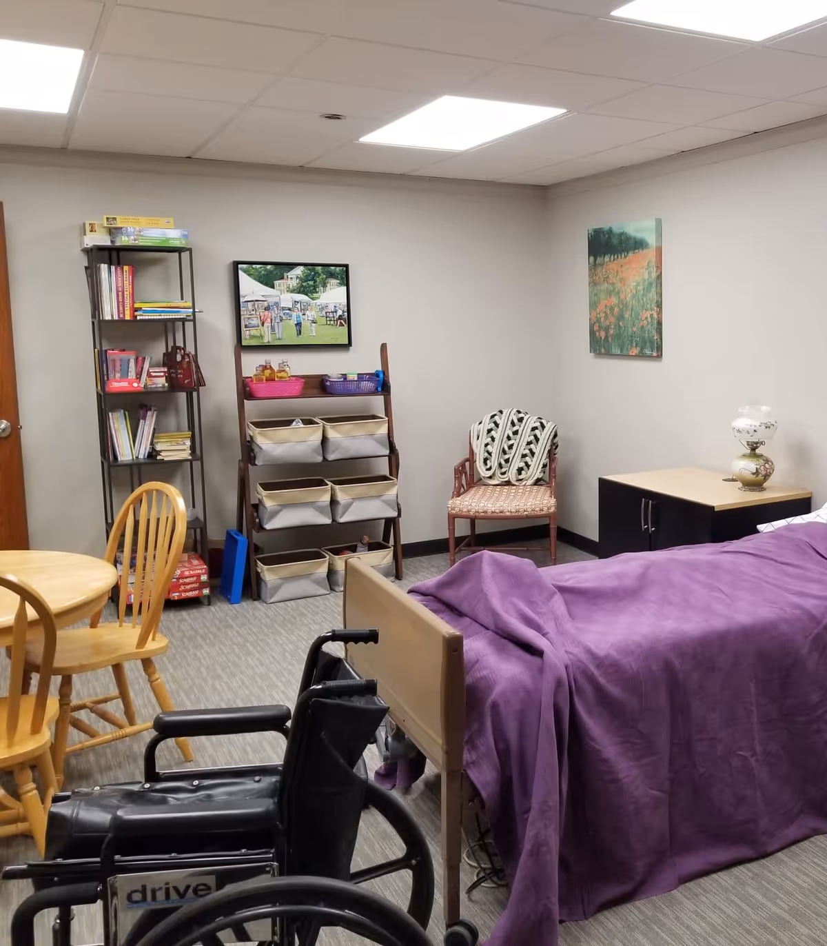 A care room with a purple-covered bed, wheelchair, wooden table, chairs, and storage shelves.