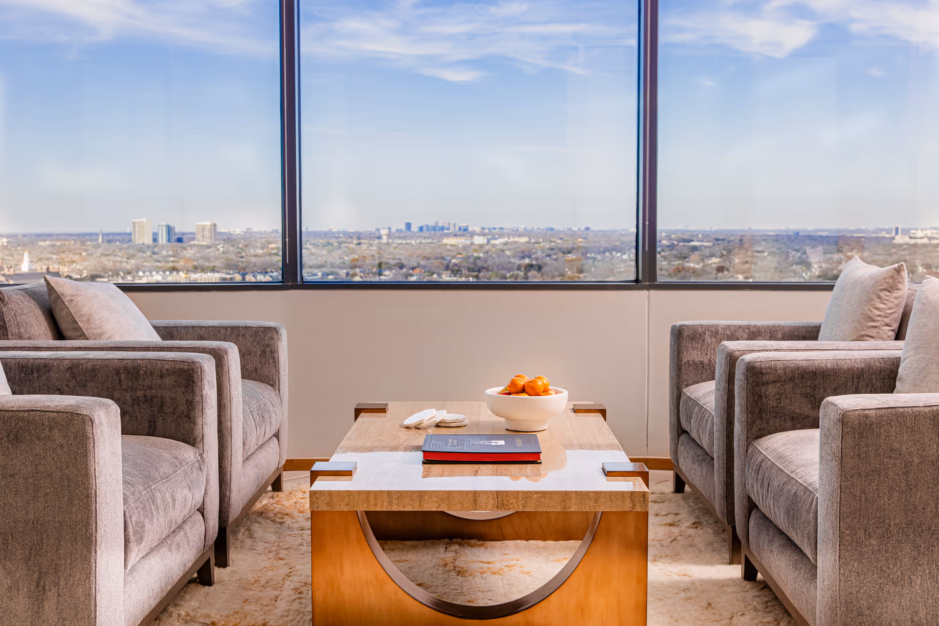 Two grey armchairs face a wooden coffee table with a bowl of oranges, city skyline beyond