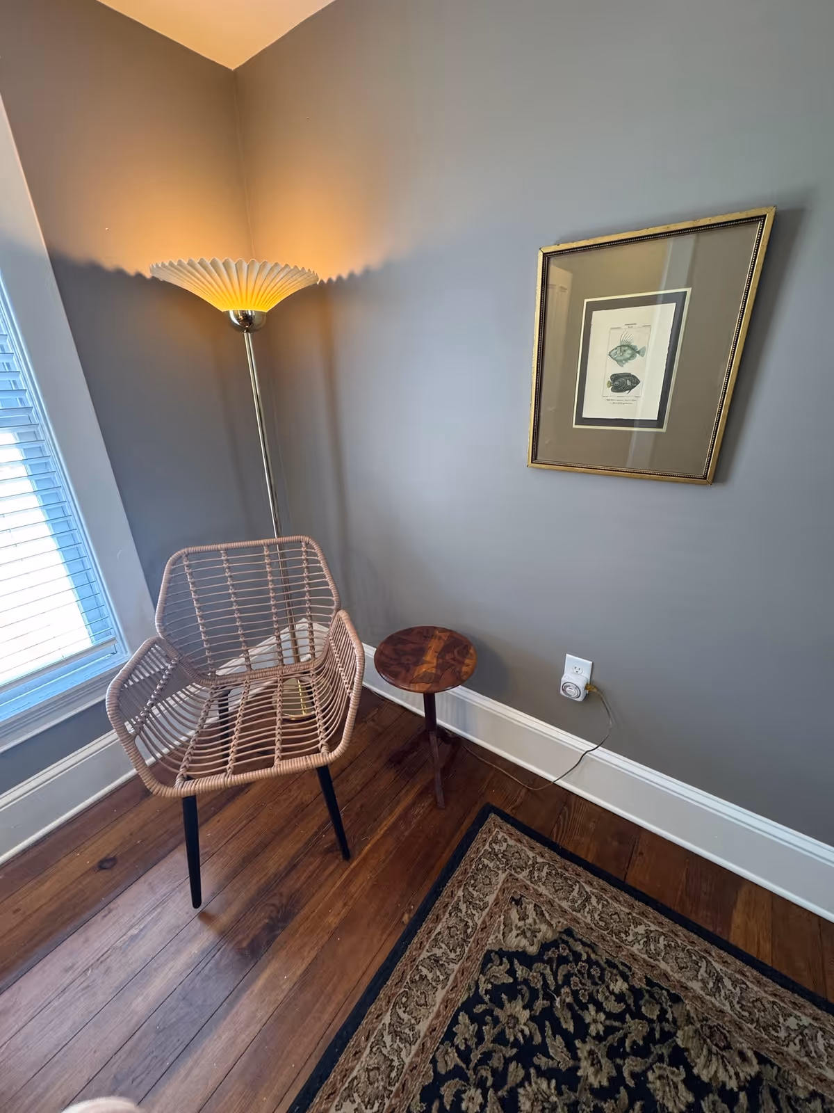 A corner of a cozy room with a wicker chair, floor lamp, framed art, and rug.