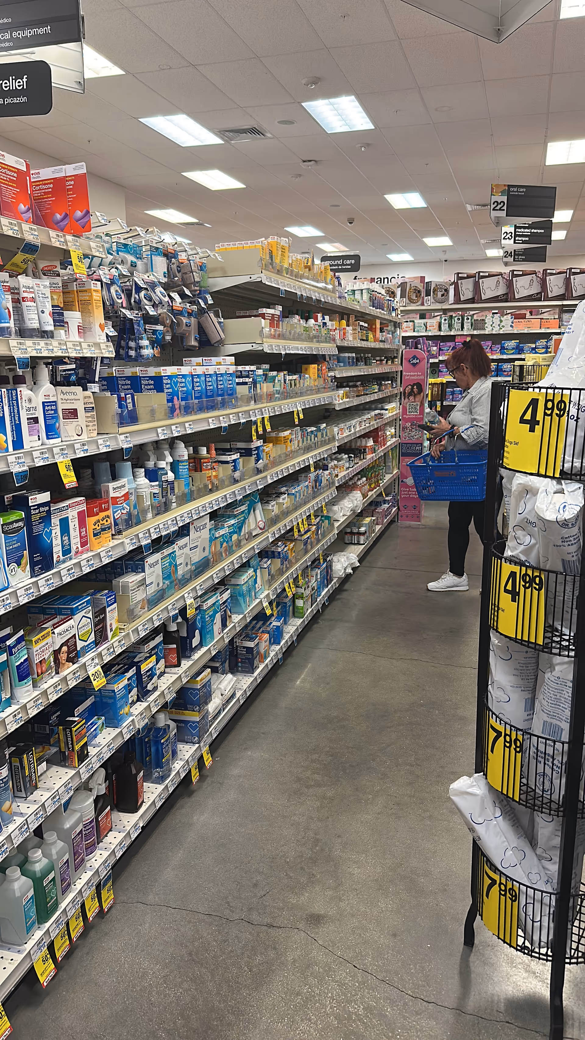 Shoppers browse a well-stocked pharmacy aisle in a store, one person with a blue basket.