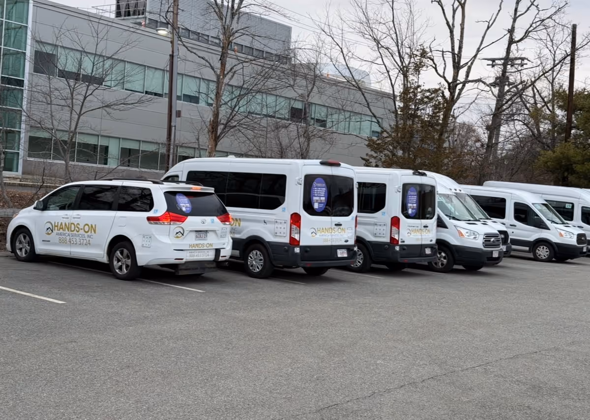 Row of white Hands-on America Services vans parked in a lot beside a modern building.