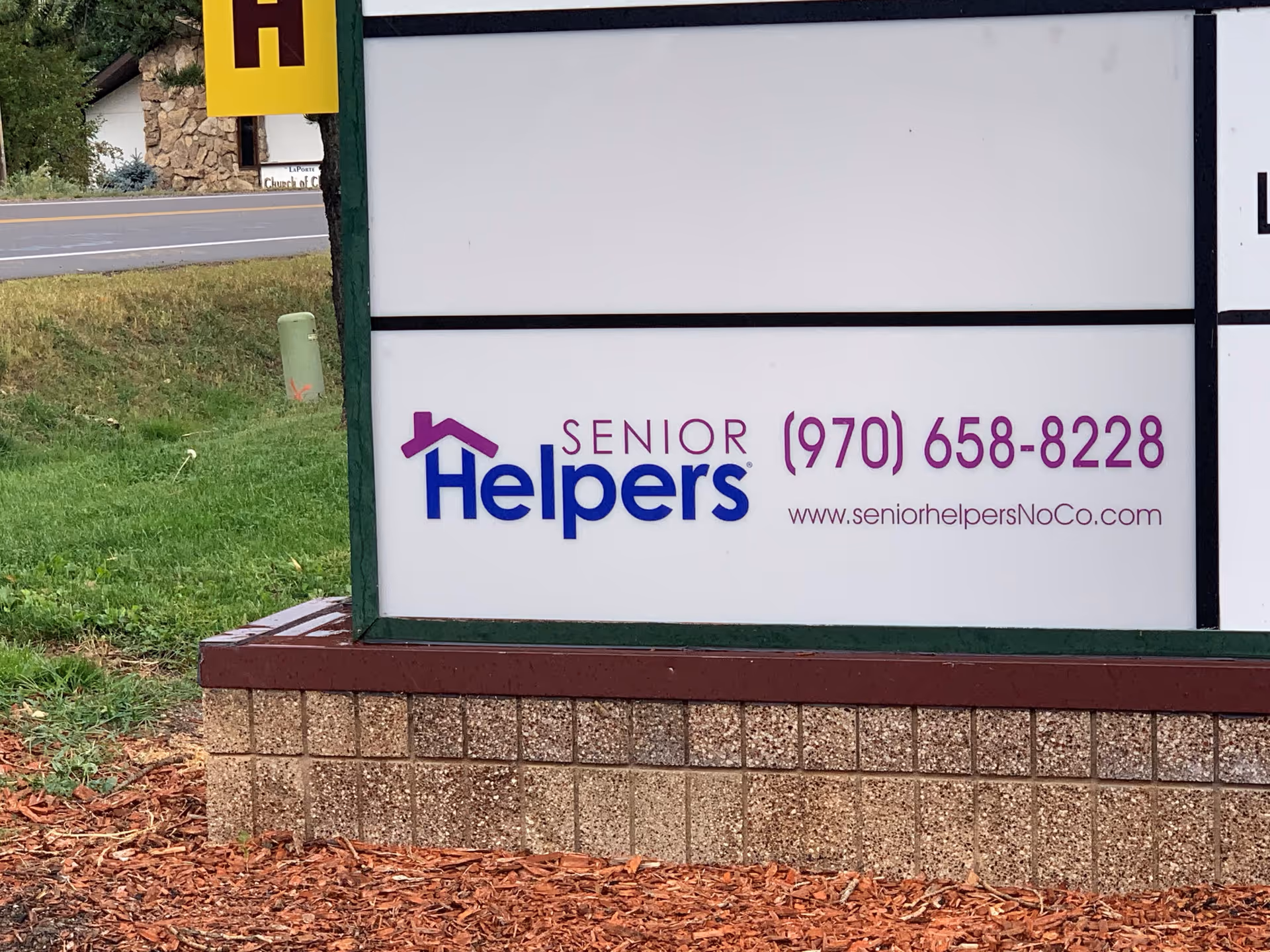 An outdoor sign displays Senior Helpers branding, phone number, and website near grassy landscaping.
