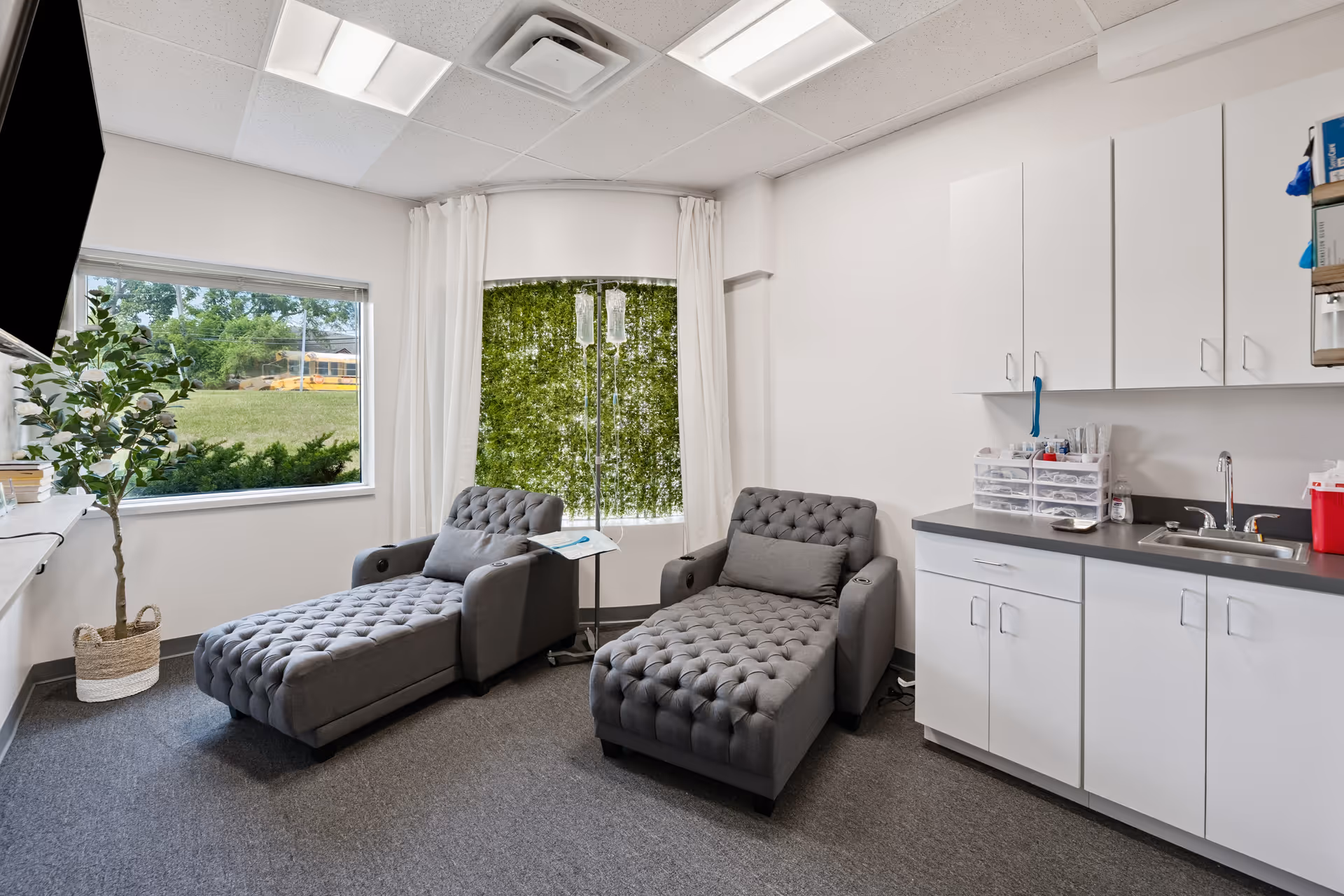 Two tufted gray lounge chairs in a bright clinic room with white cabinets and a kitchenette.