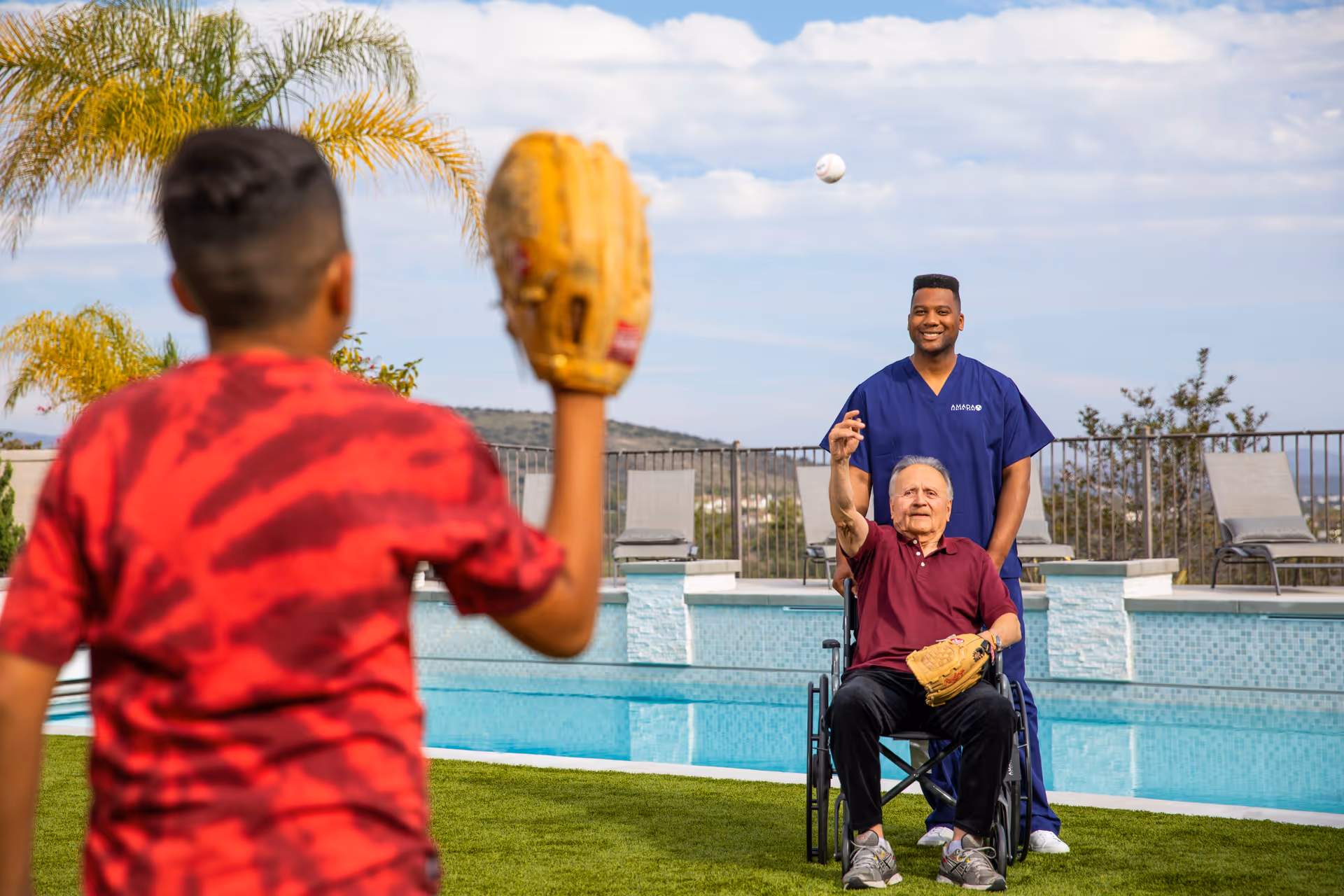 A caregiver and an elderly man in a wheelchair play catch by a pool outdoors.
