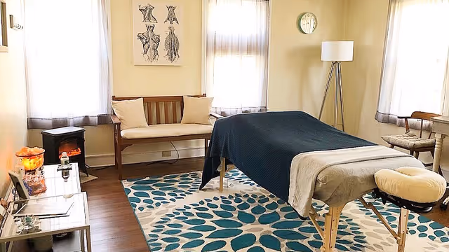 Massage room with a covered treatment table, seating, and teal-patterned rug.